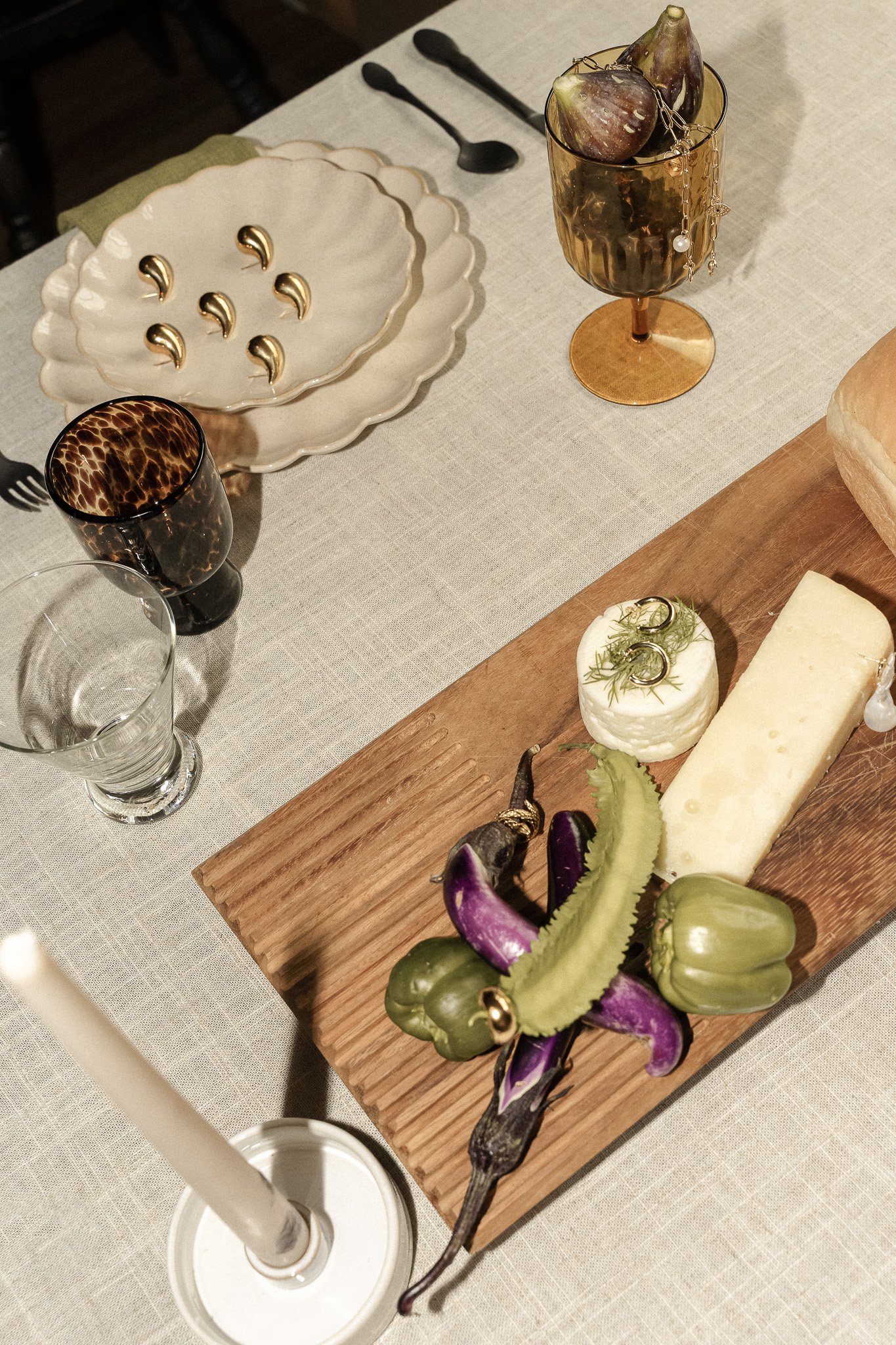 Elegant table setting with plates, glassware, and a wooden board featuring cheese, vegetables, and jewelry.