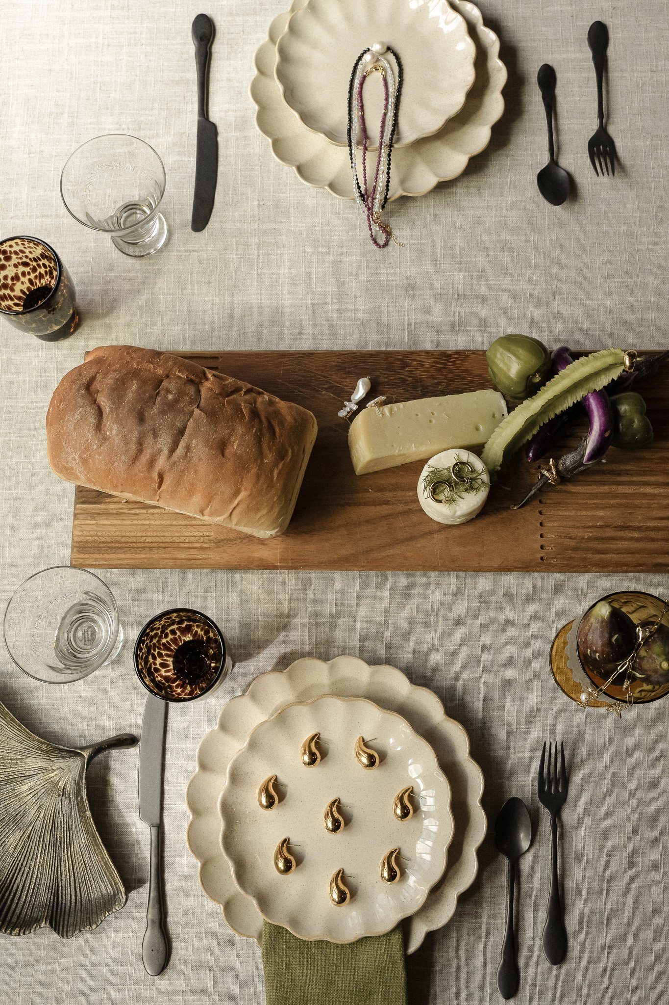 Table setting with decorative plates, loaf of bread, cheese, pickles, and black cutlery. Two glasses and a cloth napkin included.