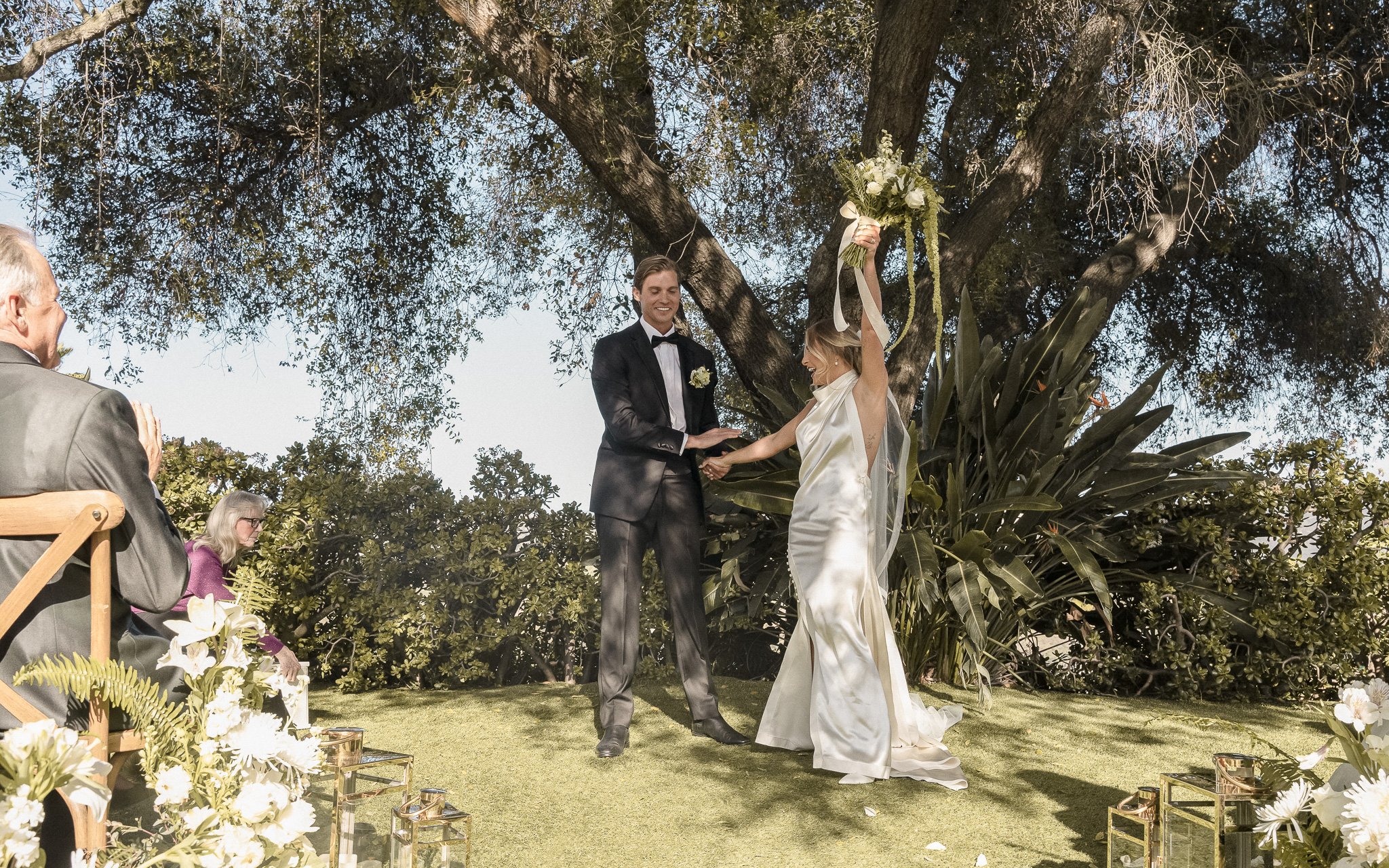 Bride and groom celebrating outdoors under trees, surrounded by guests and floral decorations.