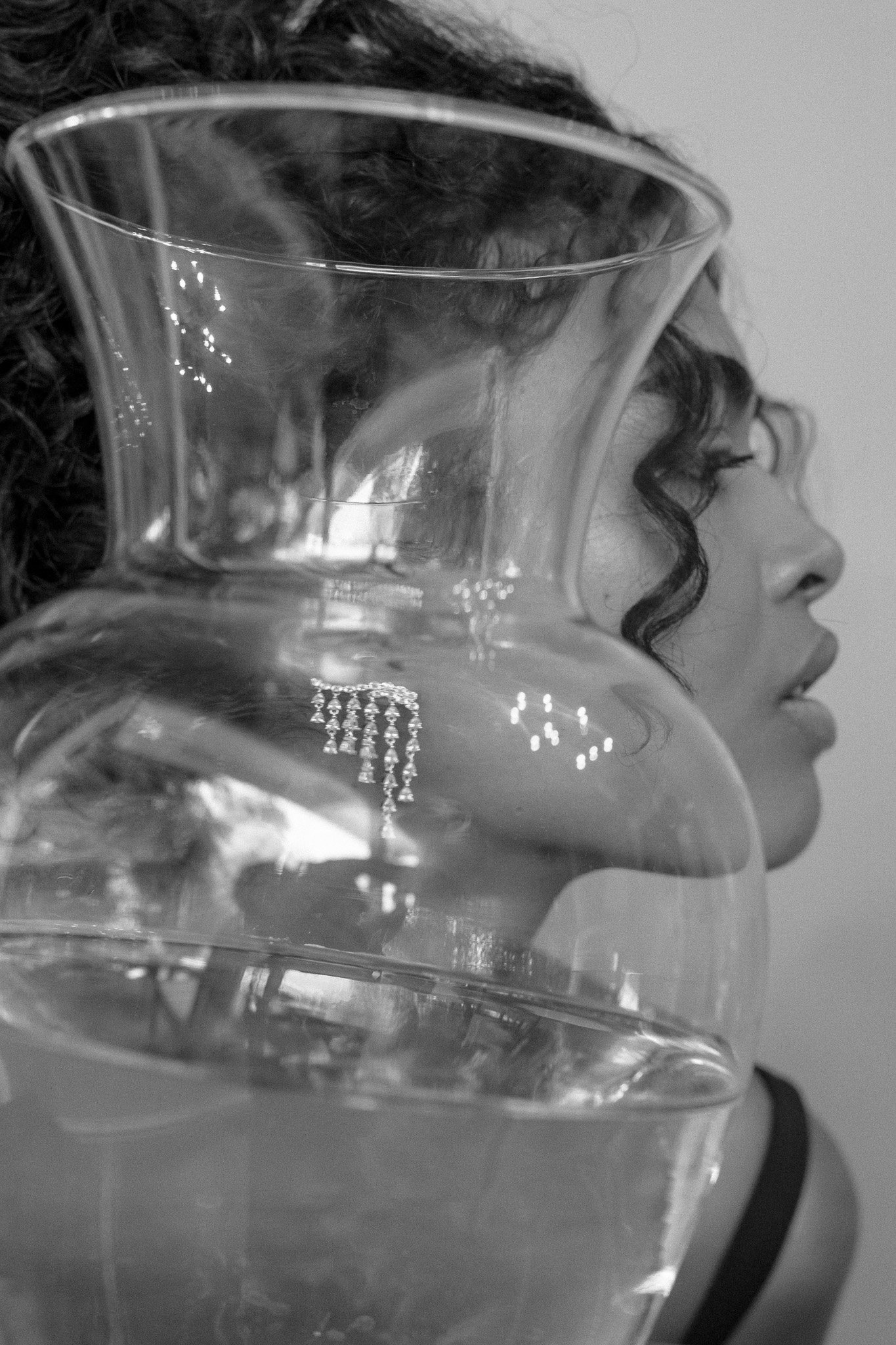 Black and white photo showing a close-up profile of a person with curly hair and earrings, partially obscured by a large glass vase.