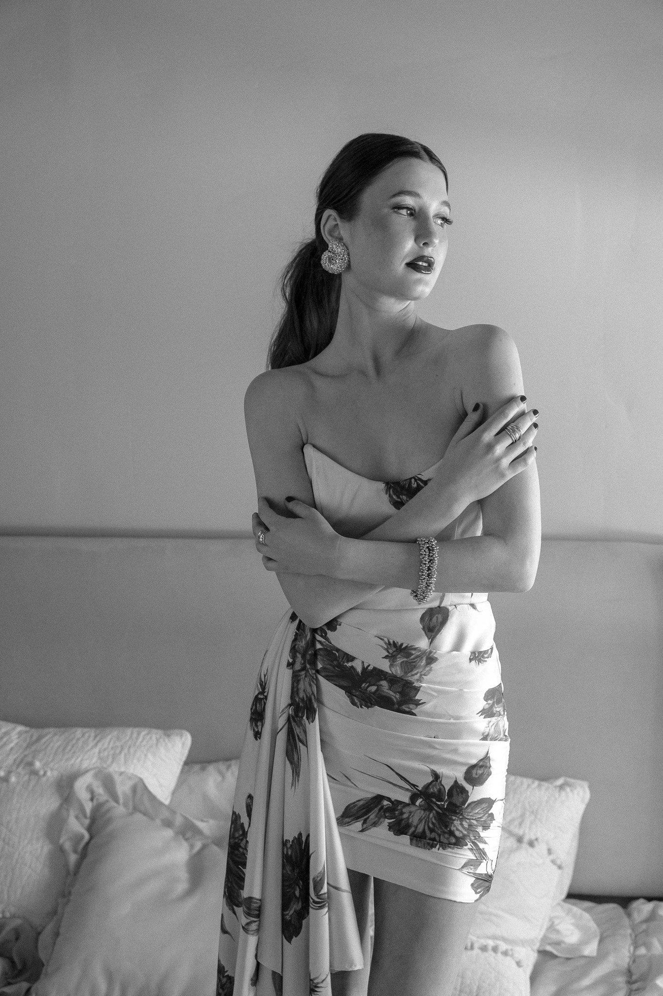 Black and white photo of a woman in a strapless floral dress, wearing large earrings and a bracelet, posing with arms crossed in front of a wall with cushions.