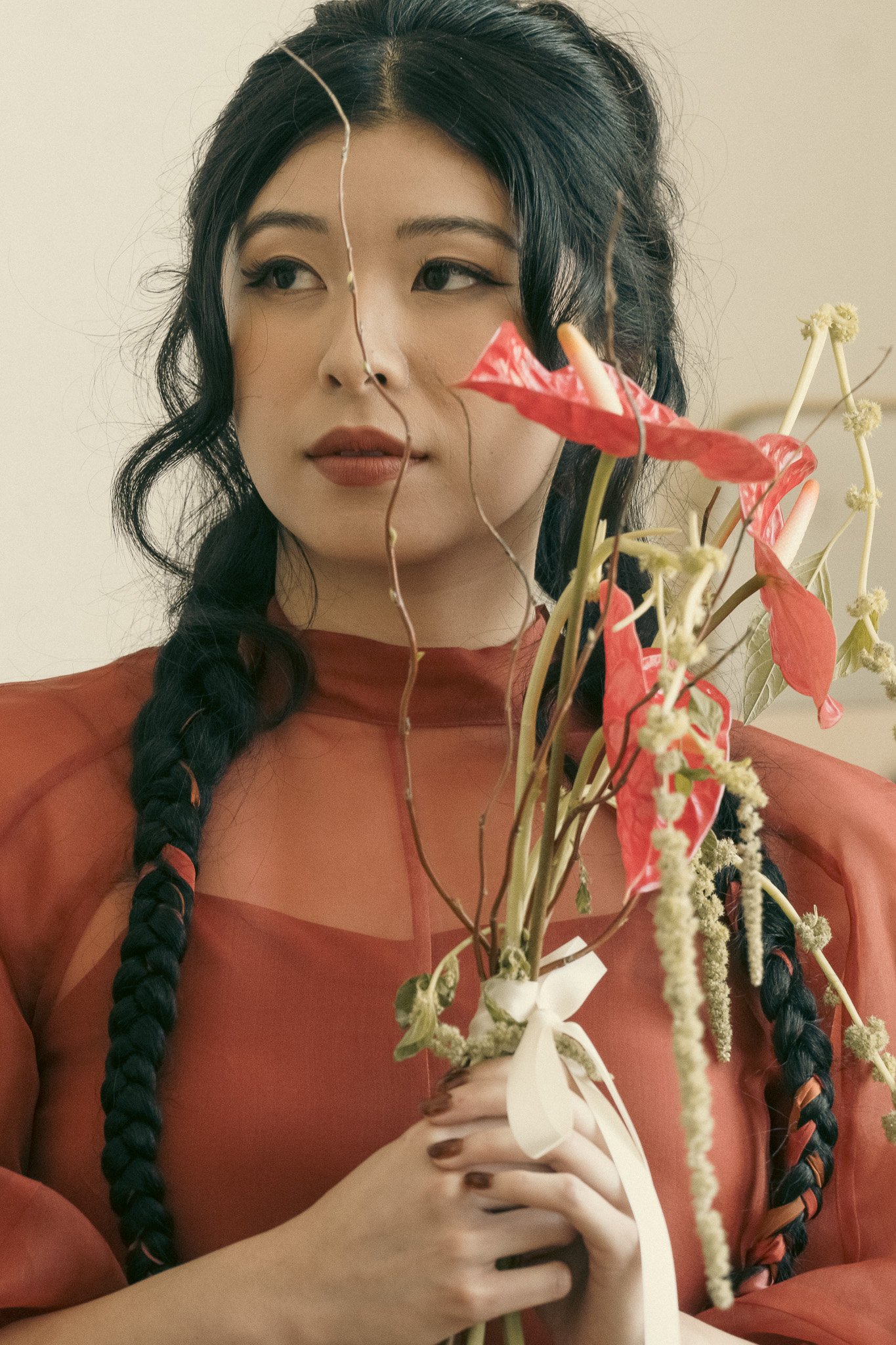 Woman with long braided hair holding bouquet of red anthurium flowers, wearing a red semi-transparent blouse.