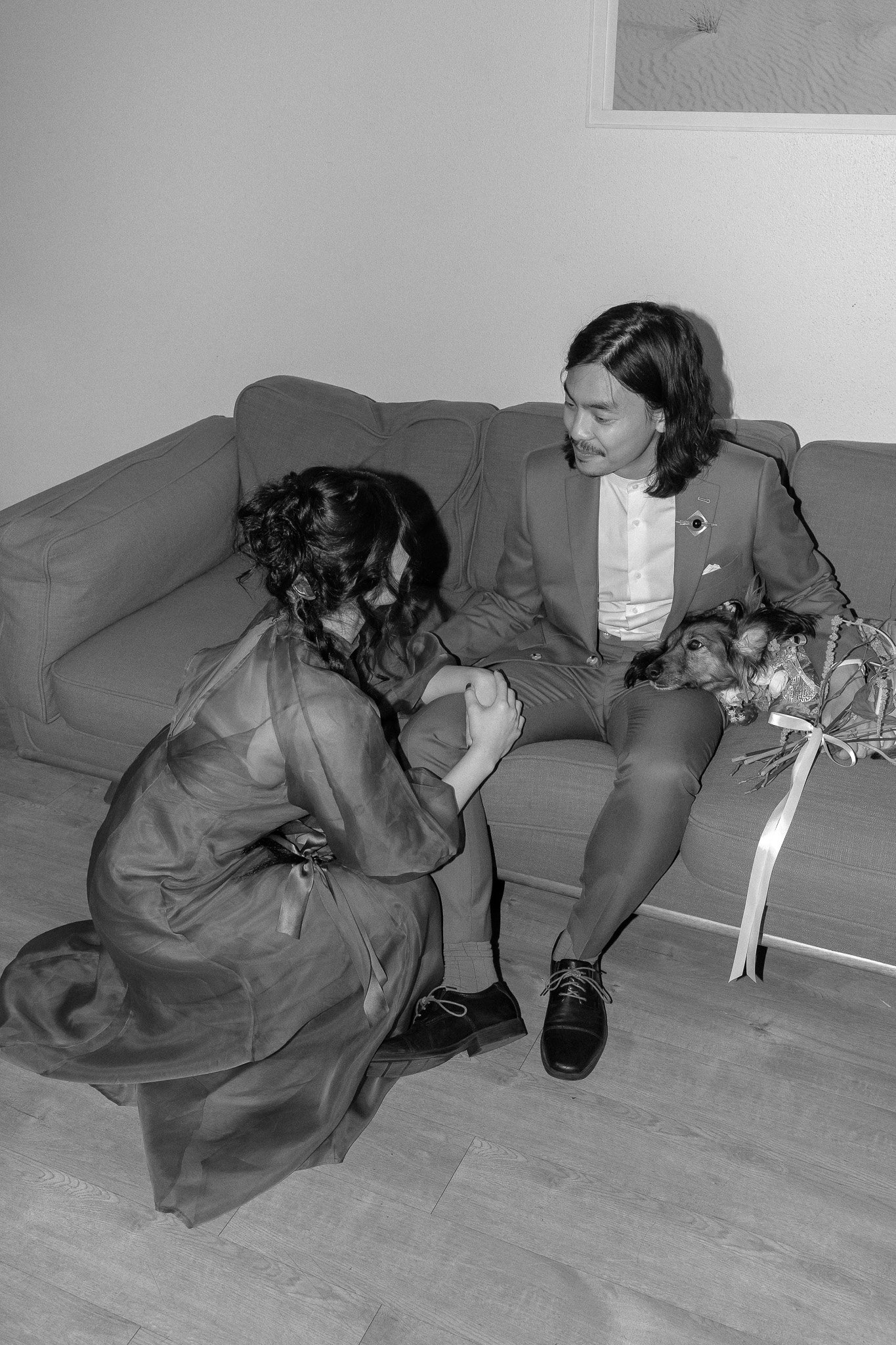 A man in formal attire and a woman in a gown are on a sofa with a dog resting on the man's lap. The woman is kneeling beside the sofa, gazing at the man. A bouquet is beside the dog in this black and white image.