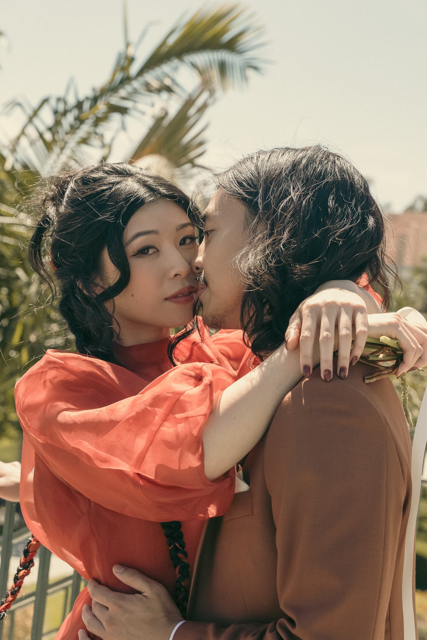 A couple embracing affectionately, with a woman in a red dress and a man in a brown suit, standing in an outdoor setting with palm trees in the background.
