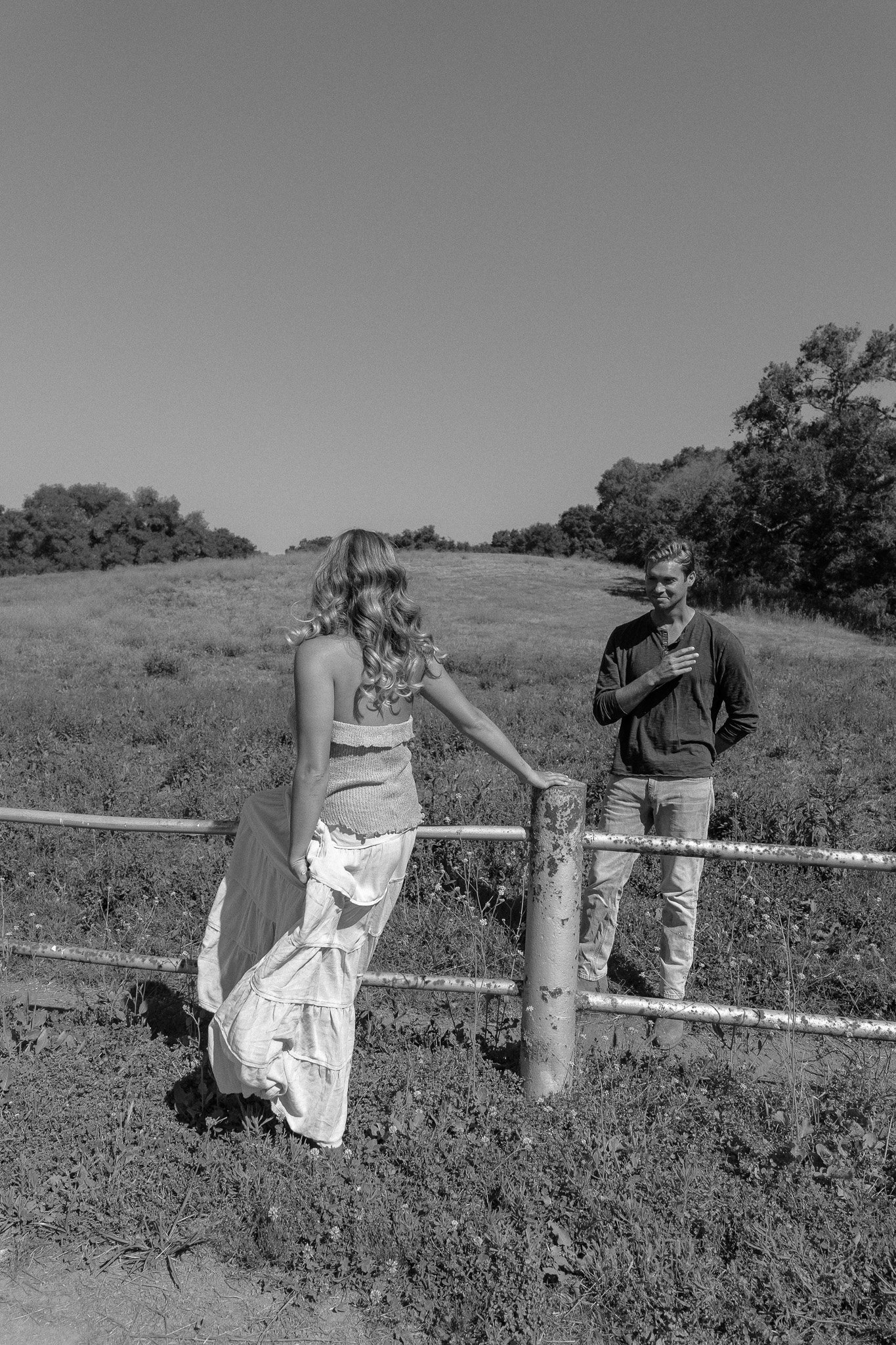 Black and white photo of a woman in a flowing dress leaning on a fence and facing a man standing on the grass in a field, with trees in the background.
