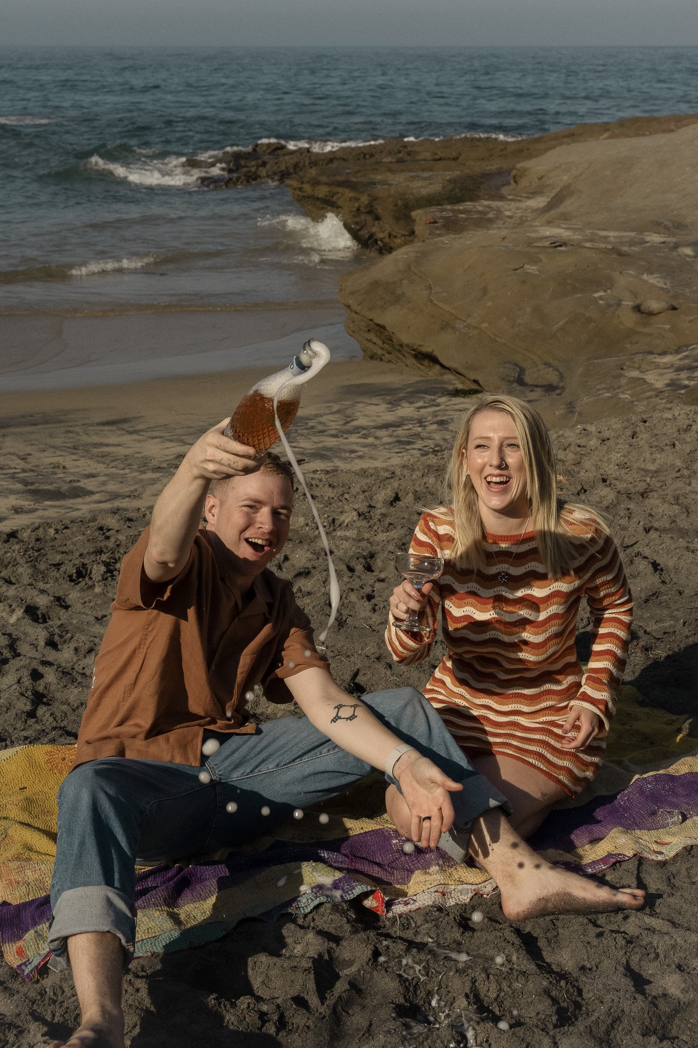 Two people sitting on a beach blanket, laughing and holding drinks, with ocean waves and rocks in the background.