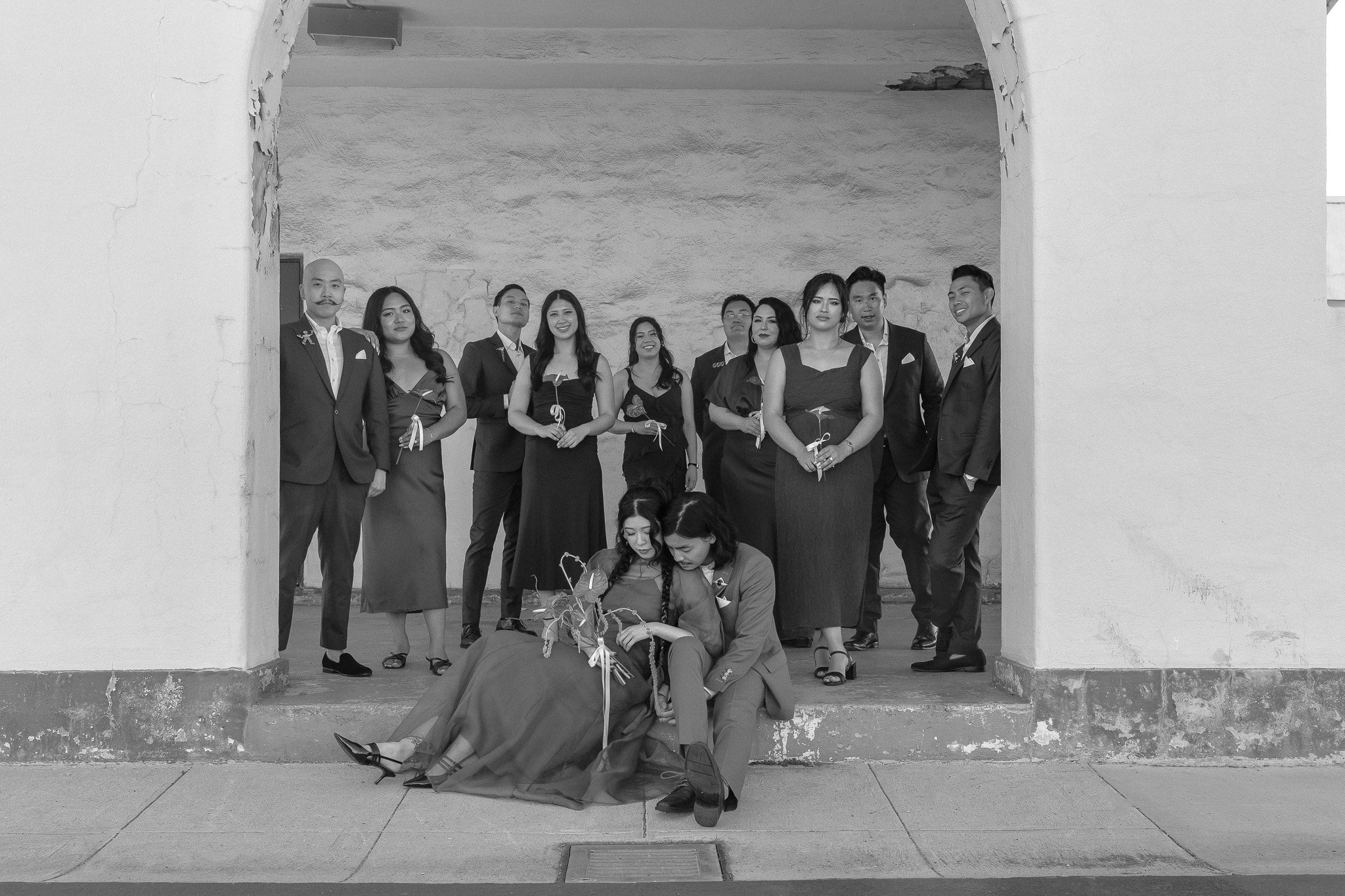Group photo of people in formal attire, with some holding flowers, gathered in an archway. A couple sits on the ground in front, with the individual on the left resting against the other.