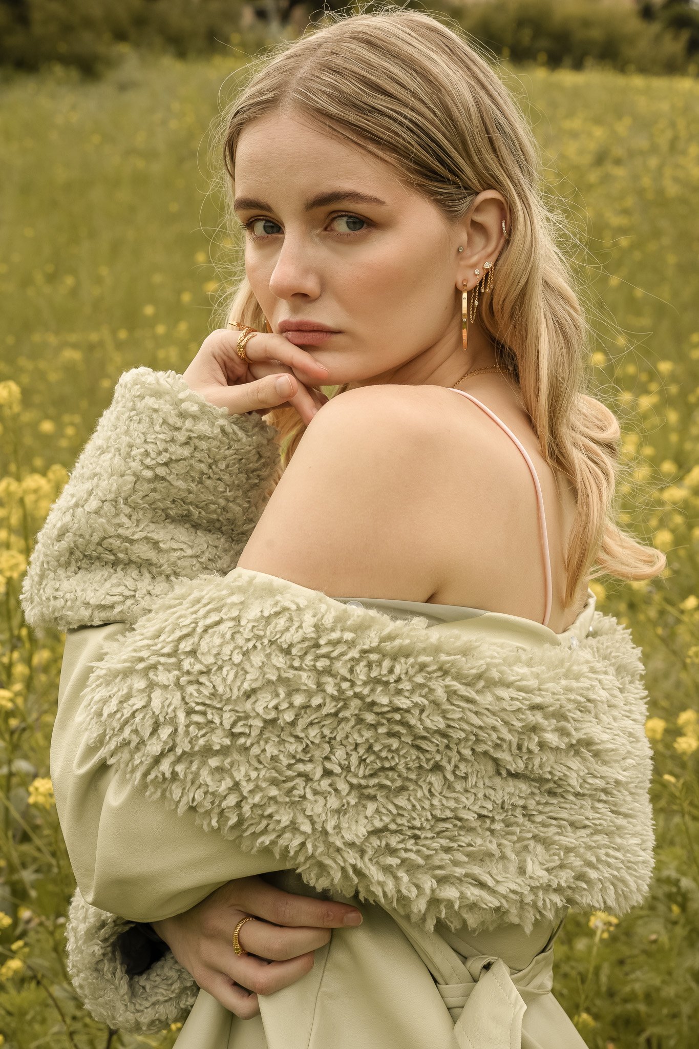 Woman with long blonde hair wearing a fluffy green coat and gold jewelry, standing in a field with yellow flowers.