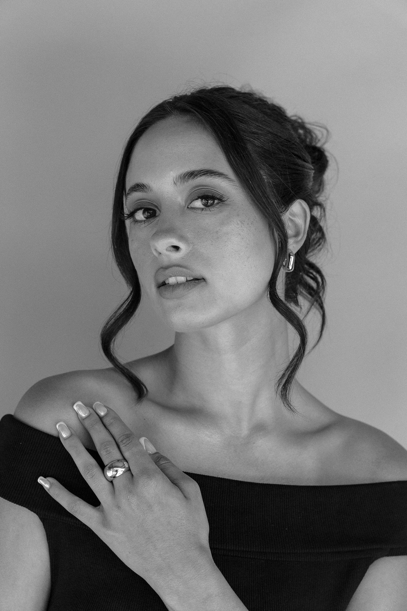Black and white portrait of a woman with tied-back hair and loose strands, wearing earrings and a ring, with a hand across her chest.