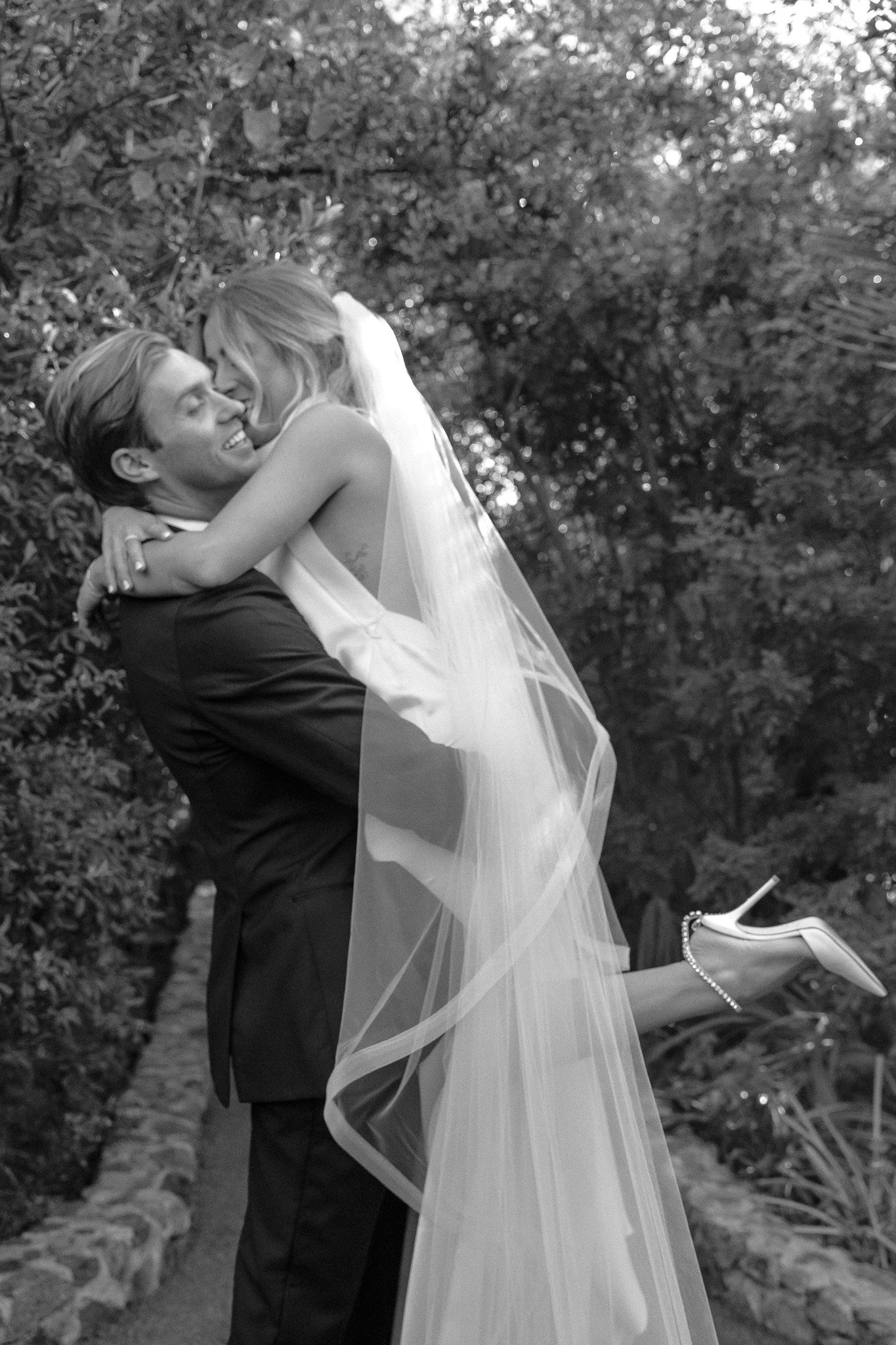 Bride being lifted by groom in an outdoor setting, wearing a veil and high heels, smiling and embracing each other.