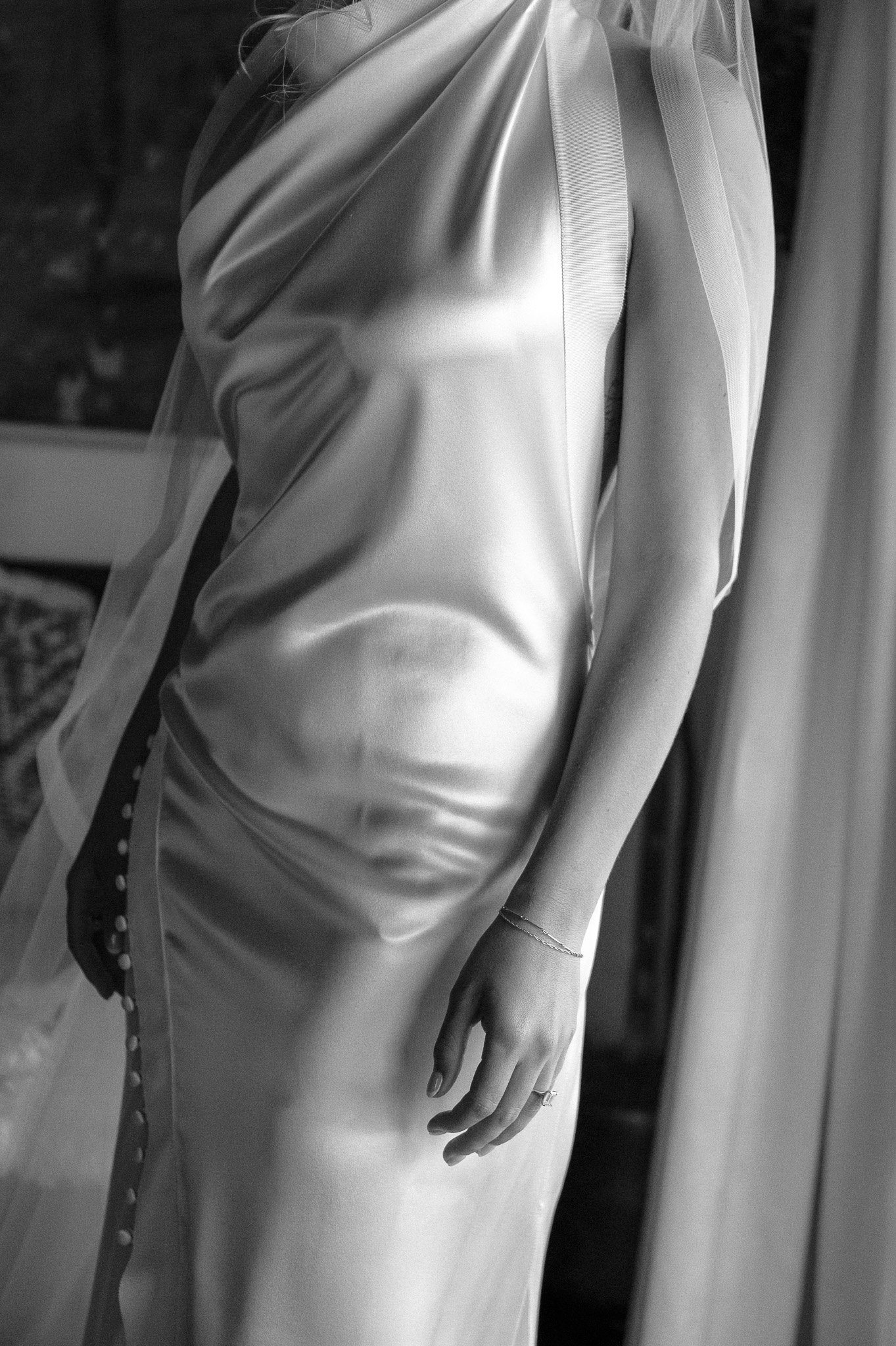 Close-up of a bride's wedding dress with satin fabric and button detail, showing part of her arm and hand wearing a bracelet and ring.