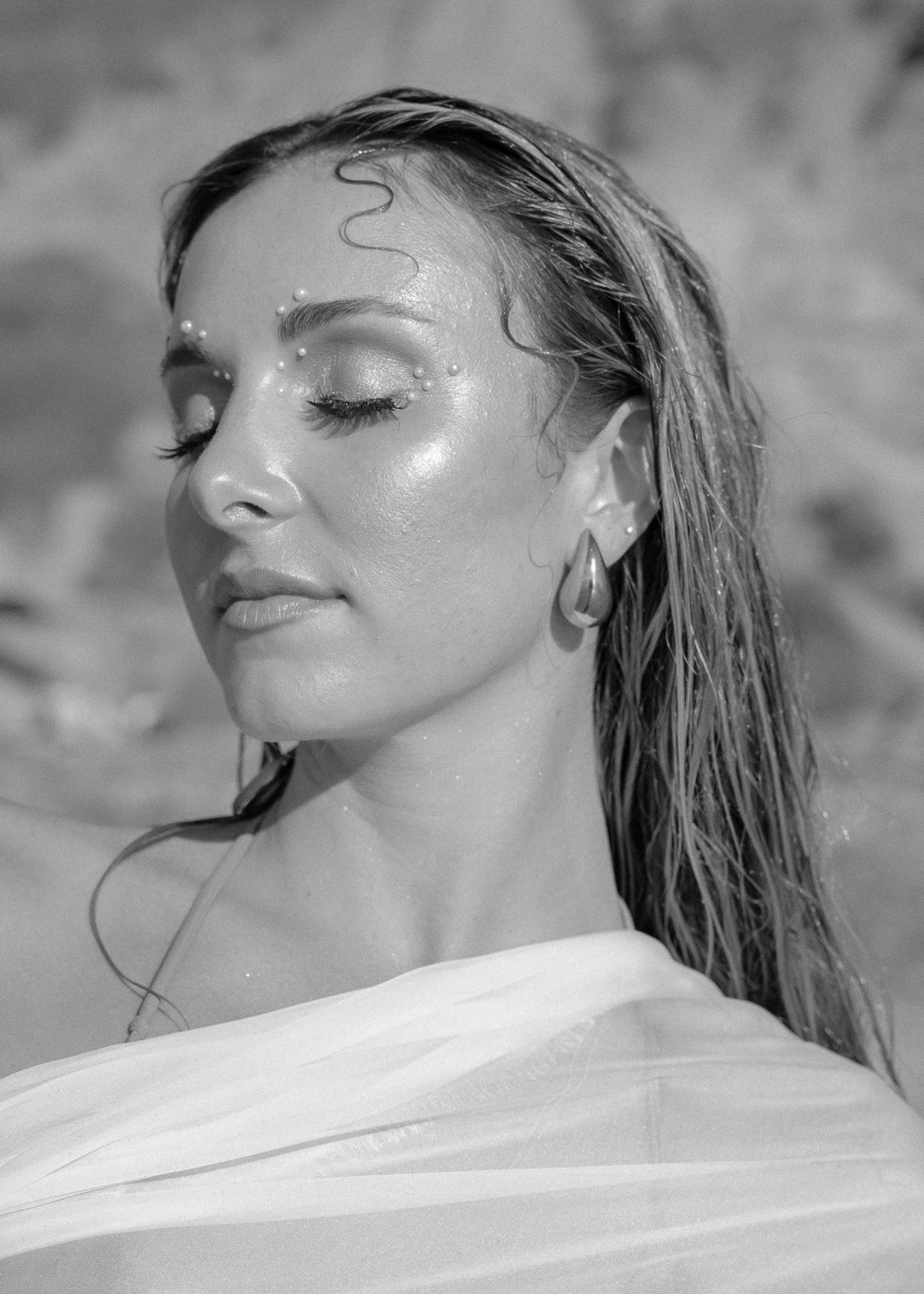 Black and white portrait of a woman with wet hair, adorned face, closed eyes, and wearing a white draped top.