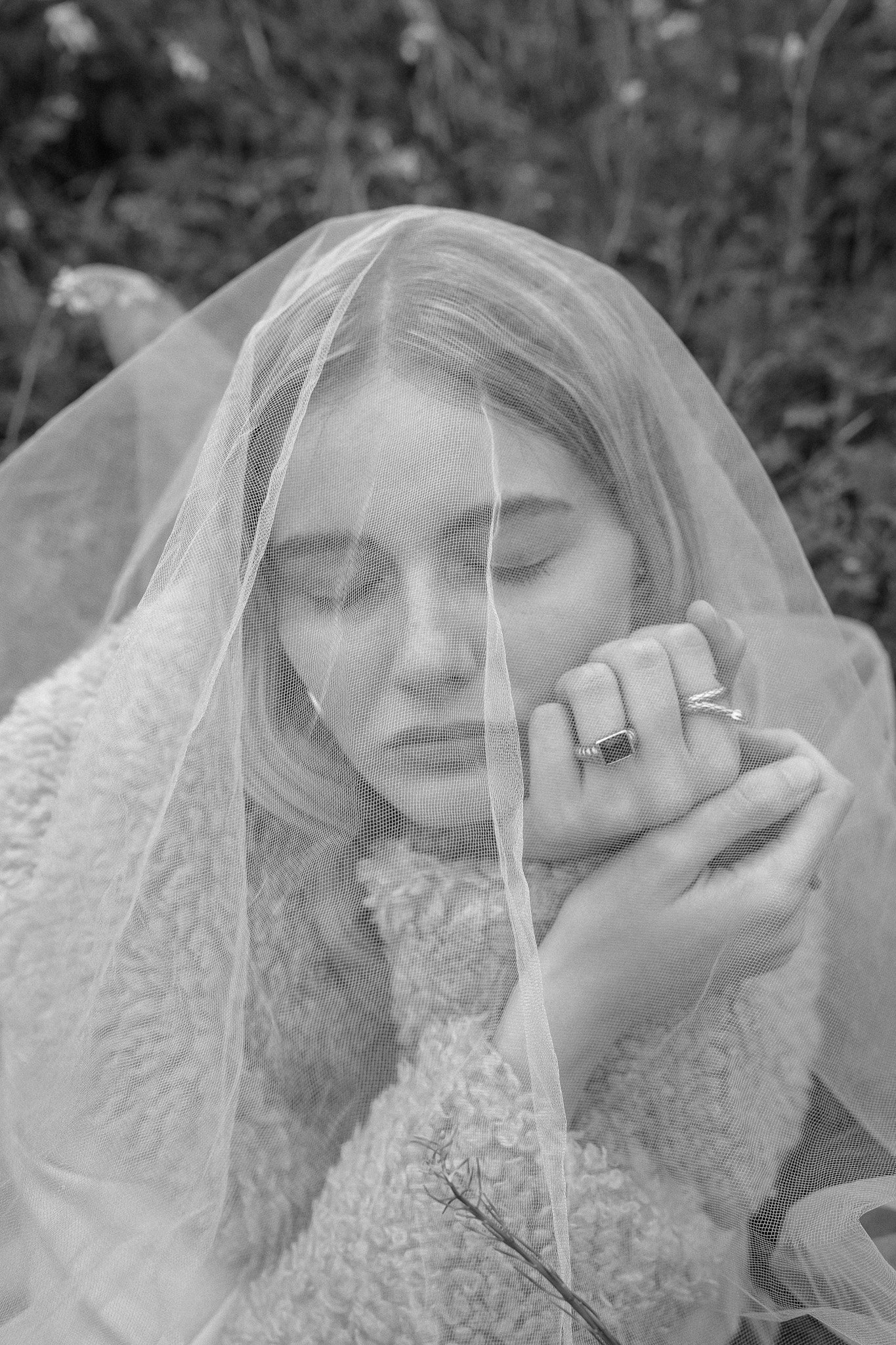 Woman with closed eyes under a sheer veil, wearing textured clothing and rings, in a natural outdoor setting.
