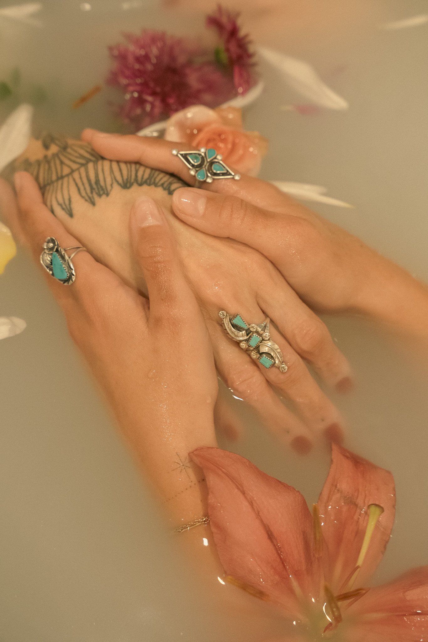 Close-up of two hands adorned with turquoise rings and a tattoo, gently holding in milky water with floating flowers.