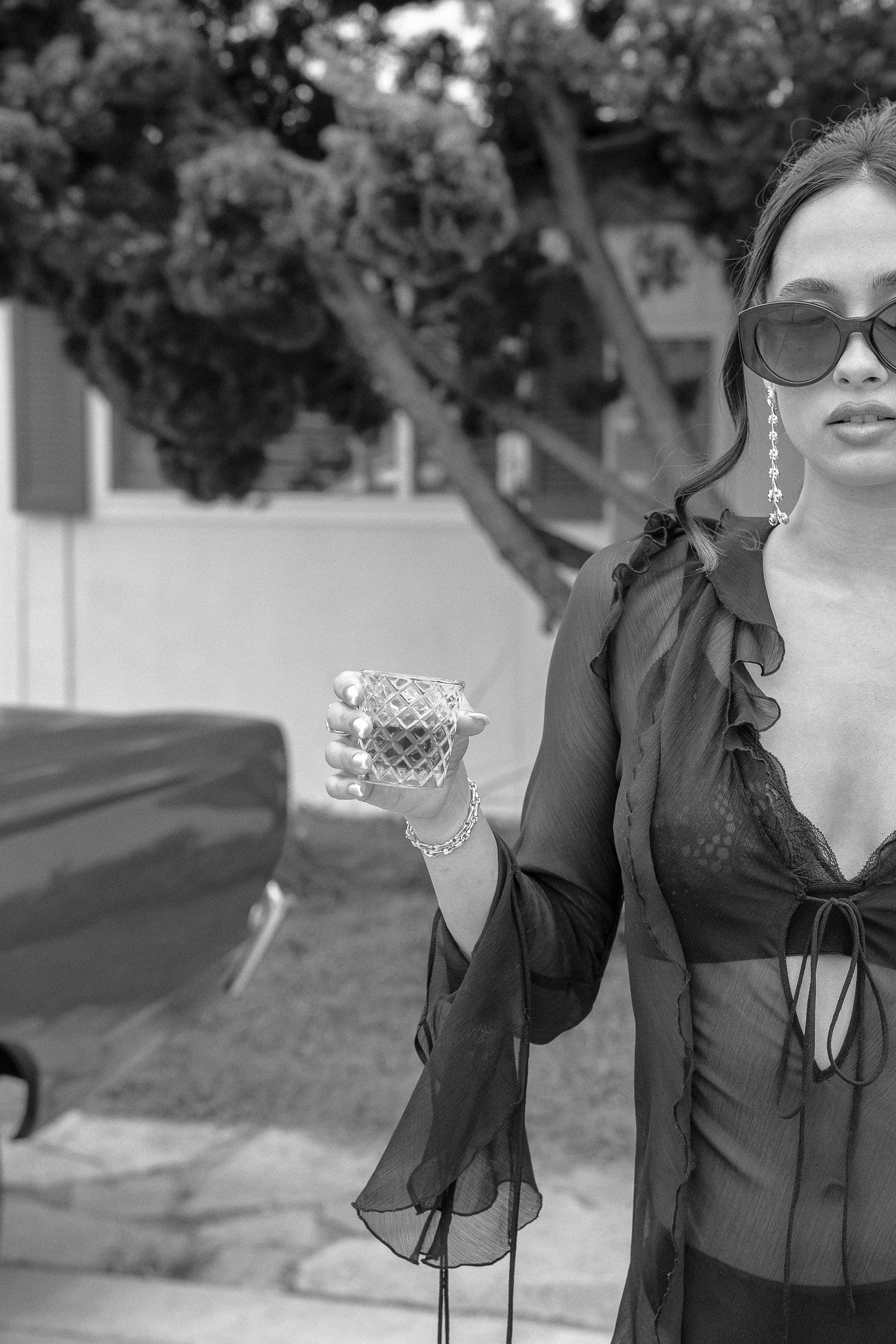 Black and white photo of a person wearing a black outfit and sunglasses, holding a textured glass, standing near a car.