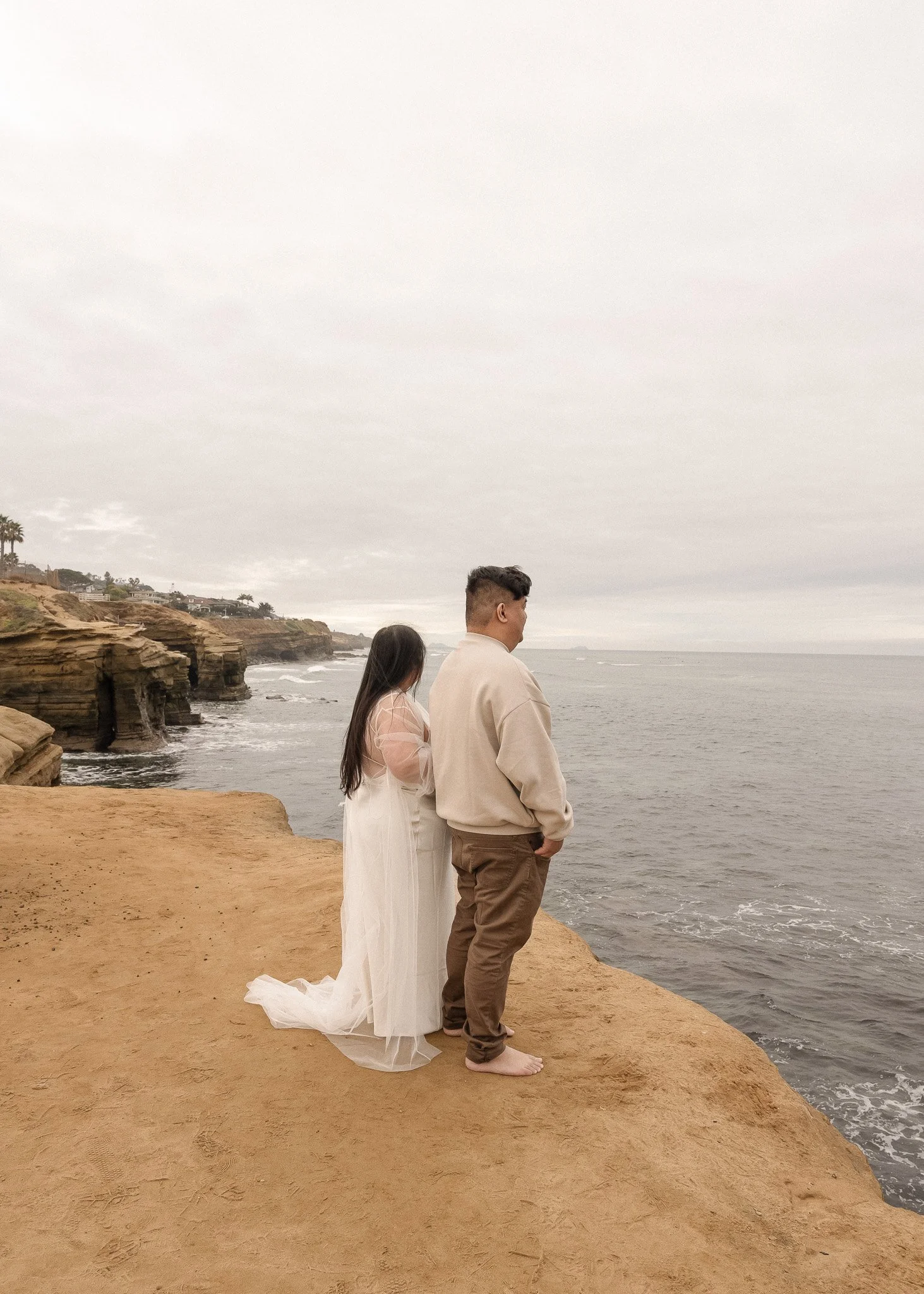 A couple standing on a rocky cliff by the ocean, facing the water. The woman is wearing a white dress and the man is wearing a beige sweater and brown pants. The sky is overcast.