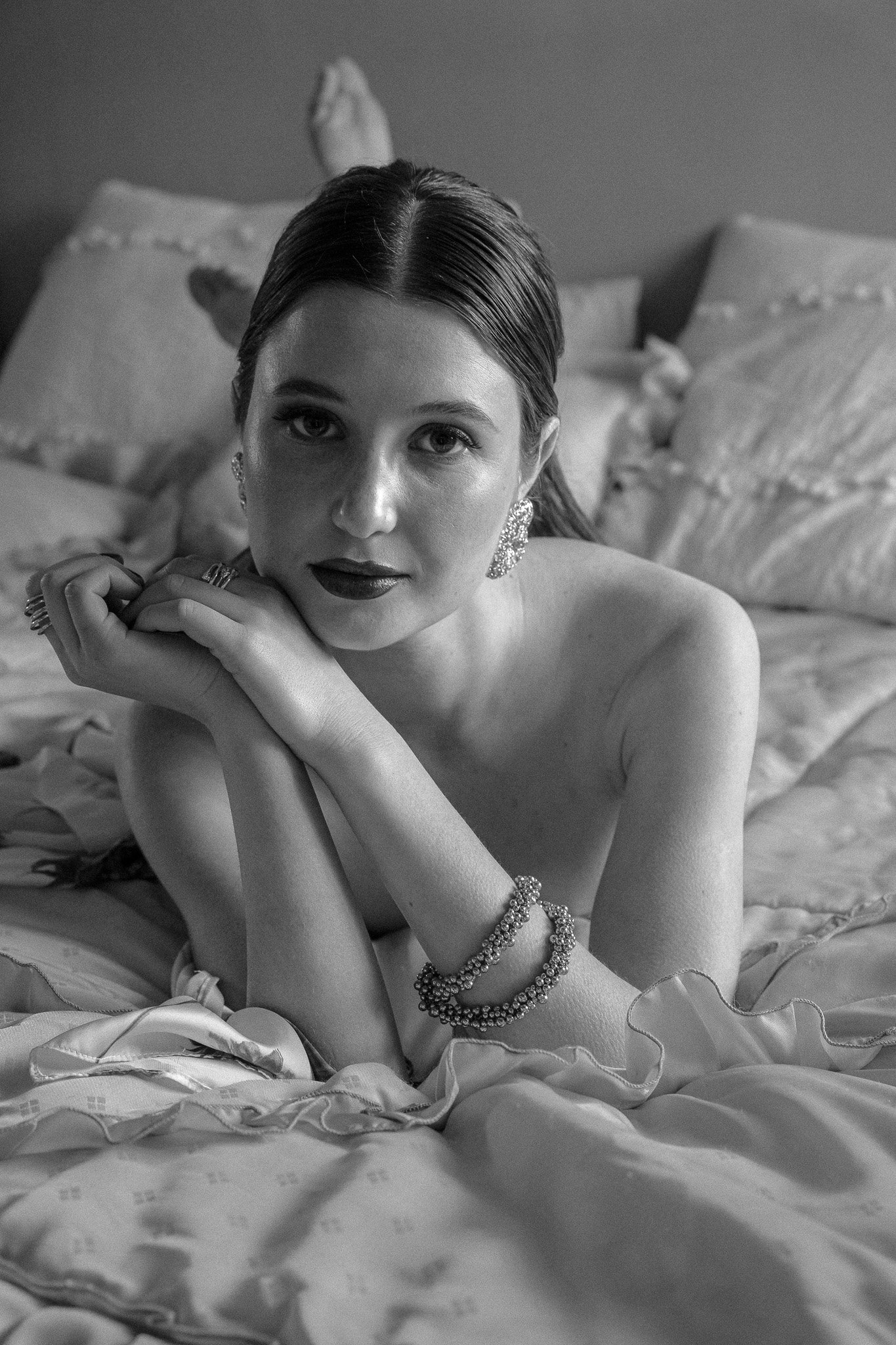 Black and white portrait of a woman lying on a bed, wearing jewelry and looking at the camera.