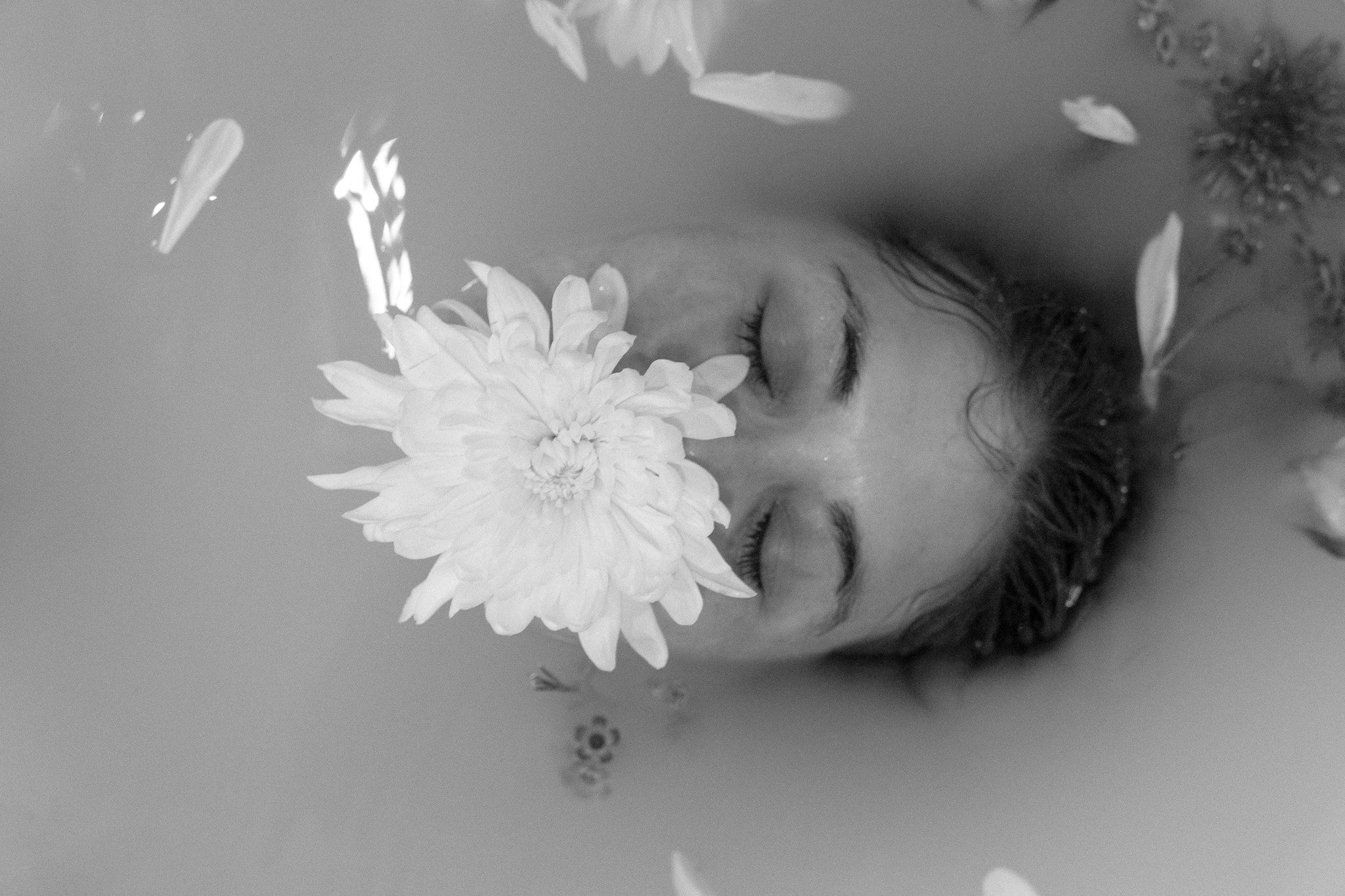 Black and white photo of a person with closed eyes submerged in milky water, with a large white flower on their face.