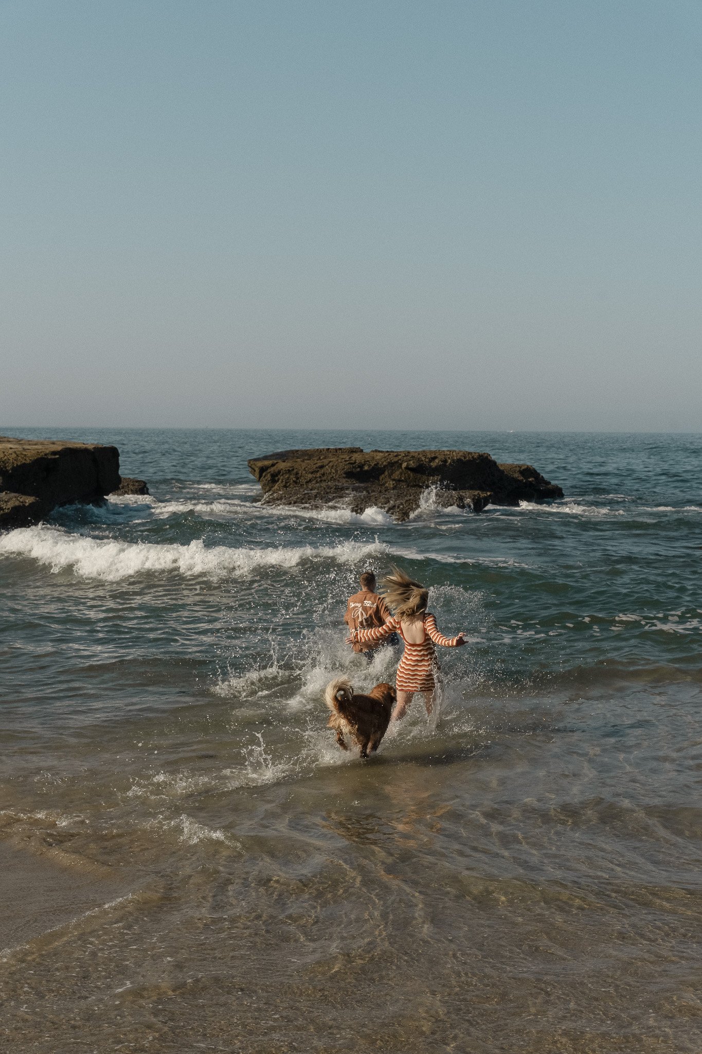 Two people and a dog running into the ocean waves near rocks on a beach.