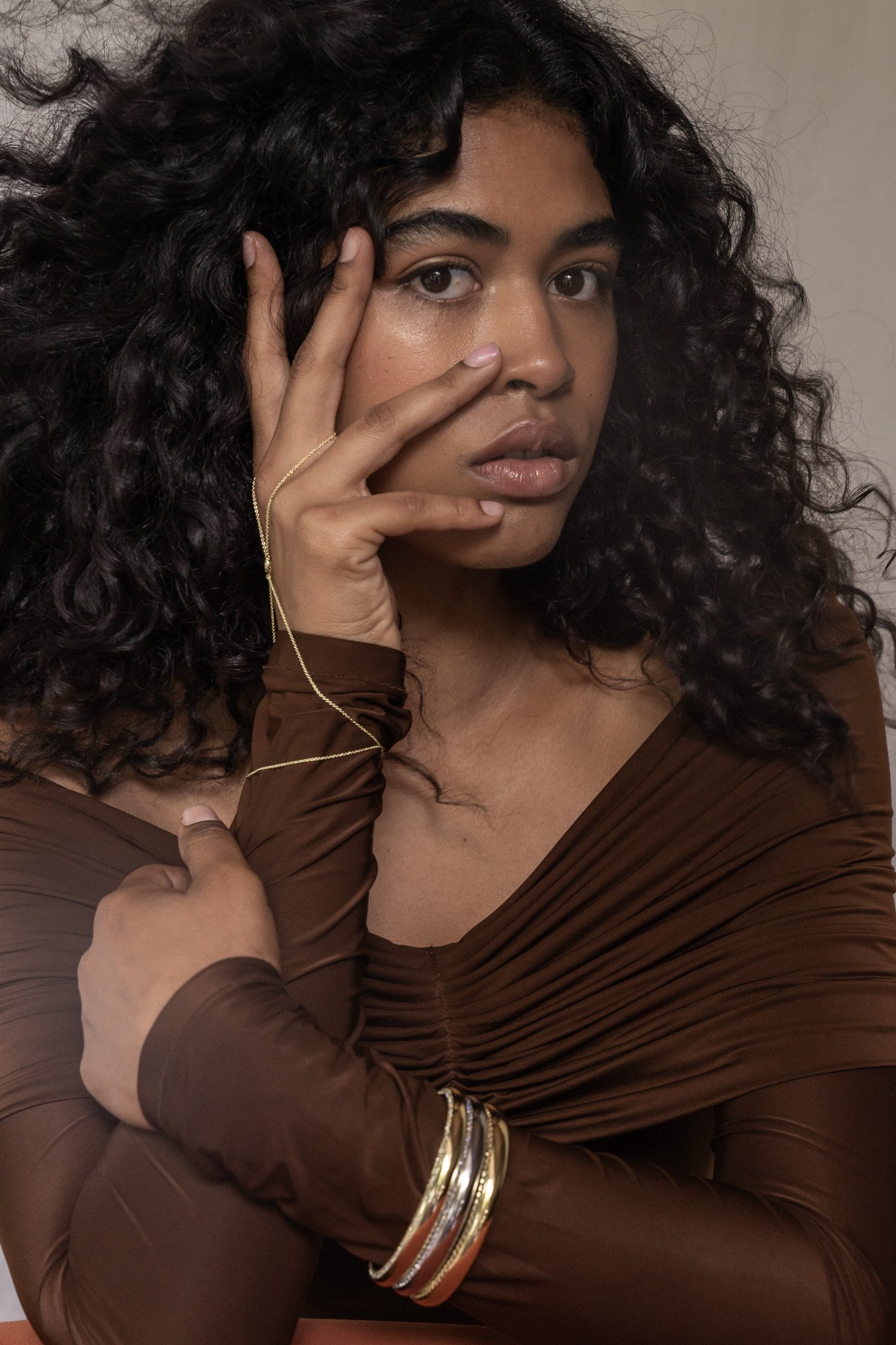 Woman with curly hair wearing a brown outfit and gold accessories, posing with hand on face.