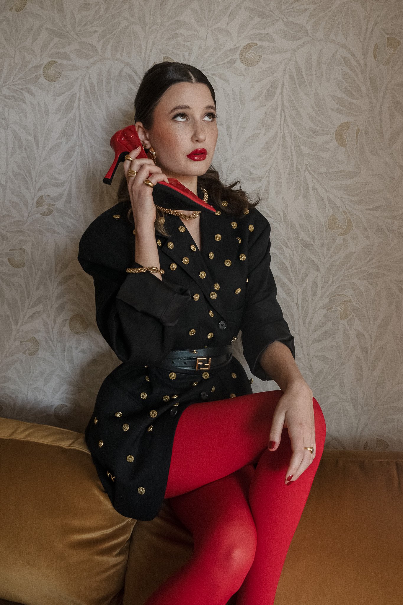 A woman with dark hair sitting on a couch, wearing a black blazer with gold buttons and red tights, holding a red high-heeled shoe to her ear like a phone.