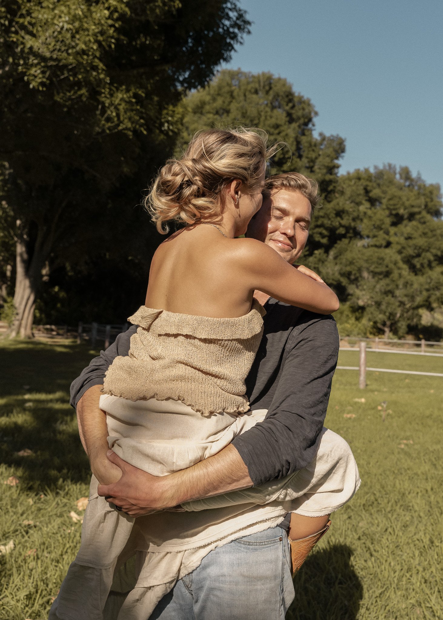 A couple embracing outdoors on a sunny day, with trees and grass in the background.