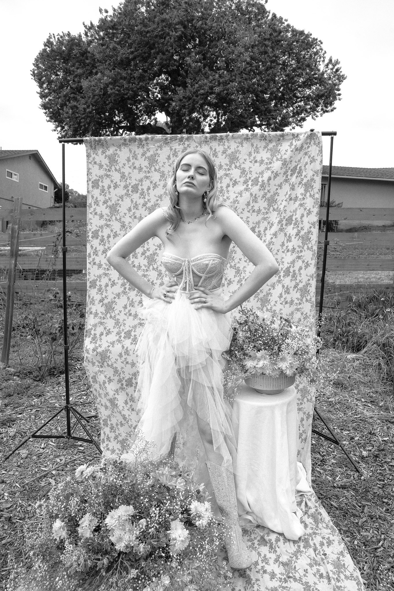 Black and white photo of a woman posing outdoors against a floral backdrop, wearing a strapless, tulle gown. She stands with hands on hips, eyes closed. Floral arrangements surround her.