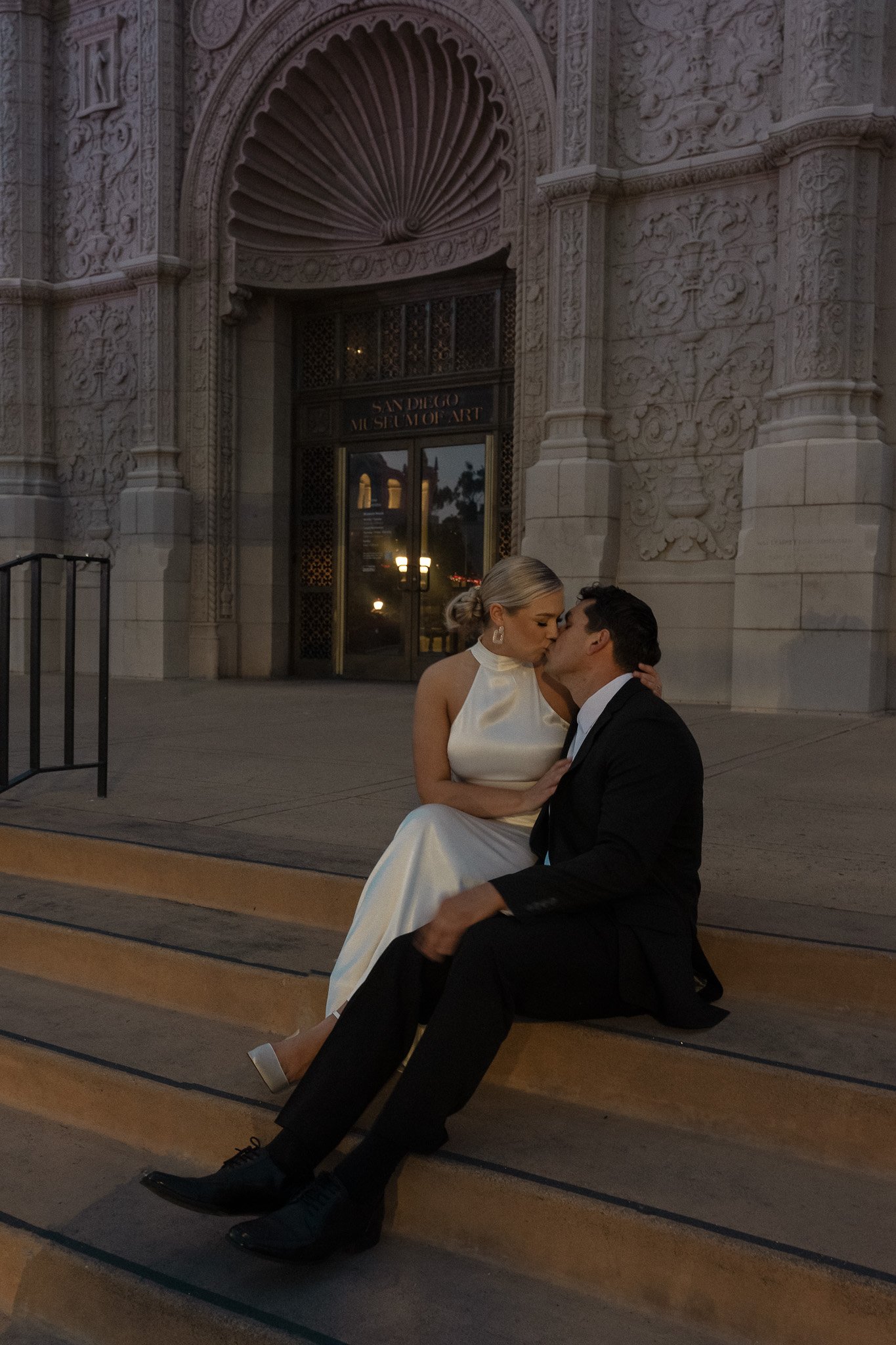 A couple sitting on the steps of a building, kissing. The woman is in a white dress, the man in a suit. The building has ornate carvings and a sign reading 'San Diego Museum of Art.'"