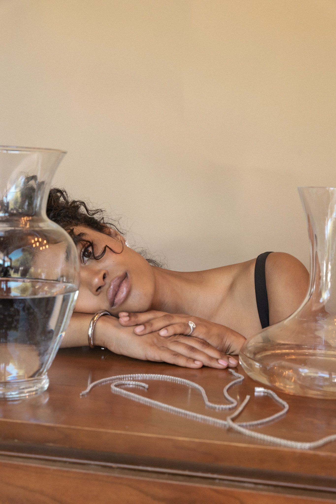 A woman with curly hair rests her head on a wooden table, surrounded by glass carafes and a silver necklace, wearing a black sleeveless top.
