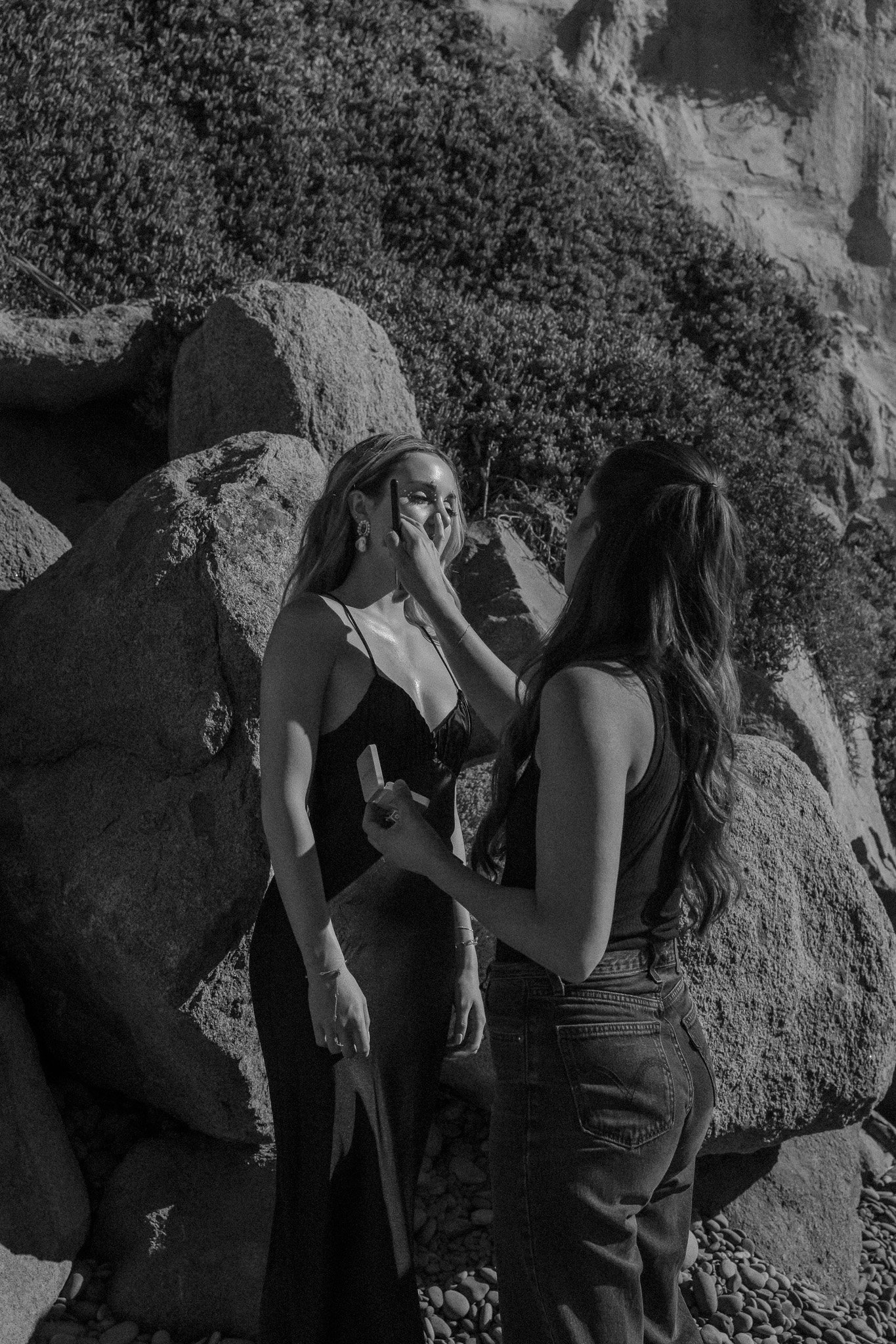 Two women by rocks and vegetation, one in a dress getting makeup applied, in black and white.