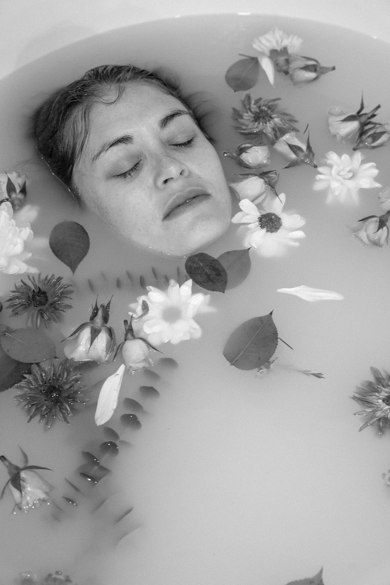 Woman's face in a milky bath with floating flowers and leaves, black and white photo.