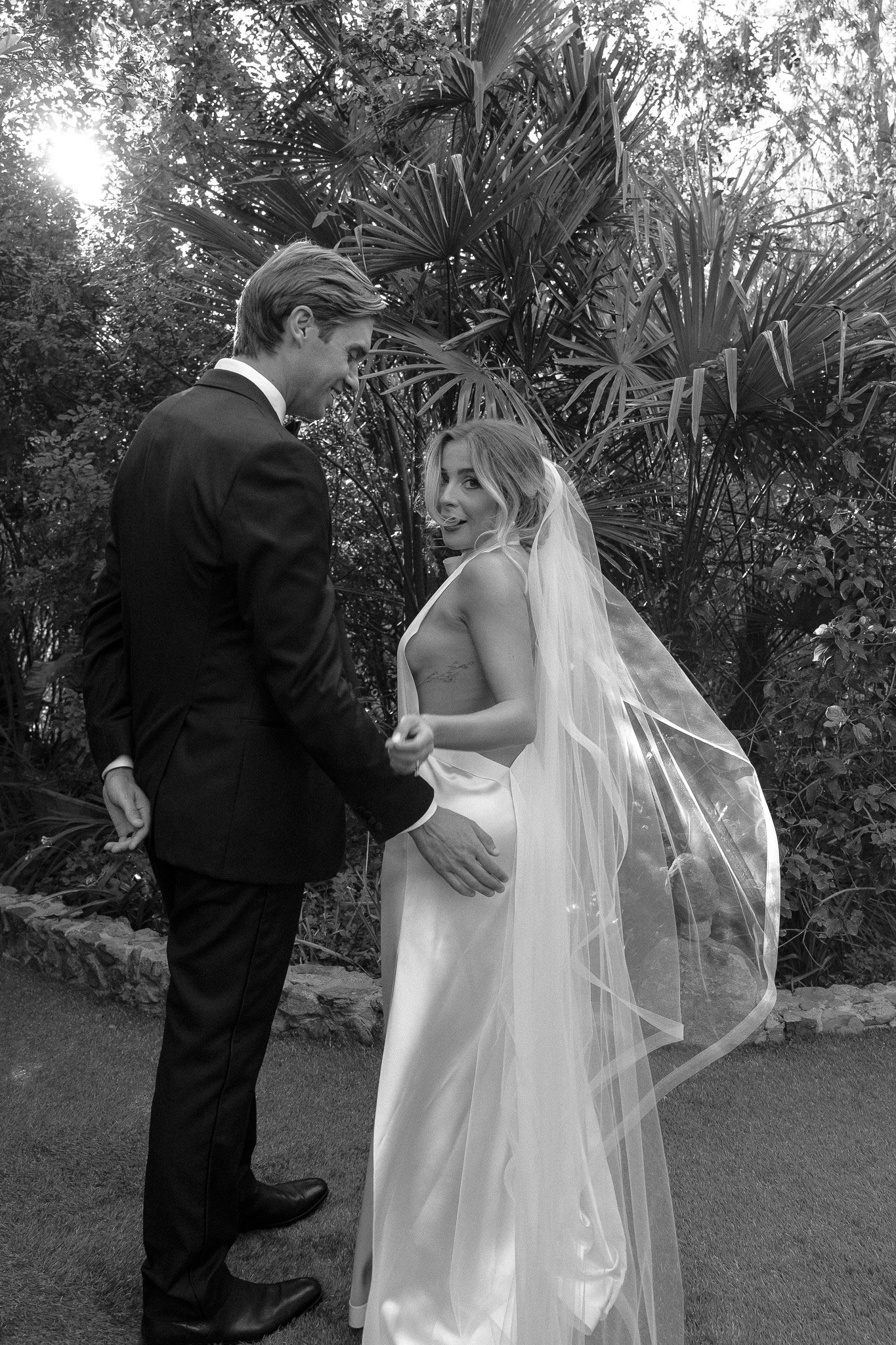 Bride and groom standing outdoors in a garden, with greenery in the background; the bride is wearing a veil and a satin dress, while the groom is in a suit, both facing each other and smiling.