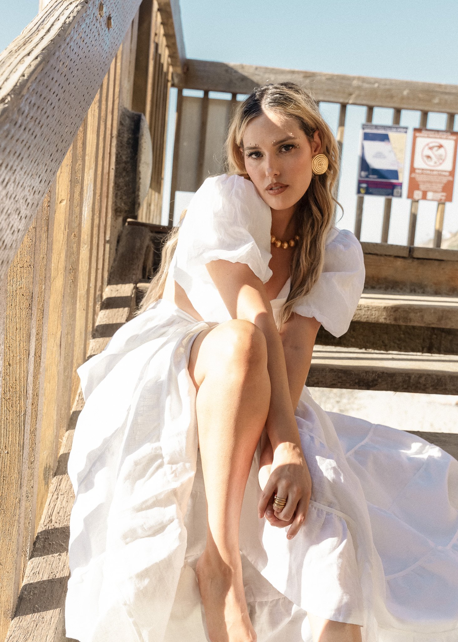 Woman in a white dress sitting on wooden steps, wearing jewelry, with a blue sky background.