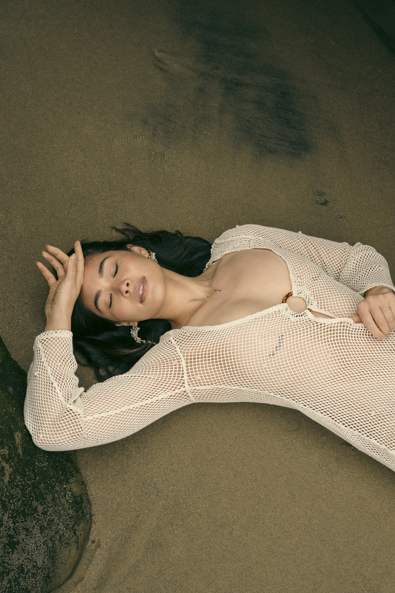 Person lying on sandy surface wearing a white mesh dress, with eyes closed and one hand on forehead.