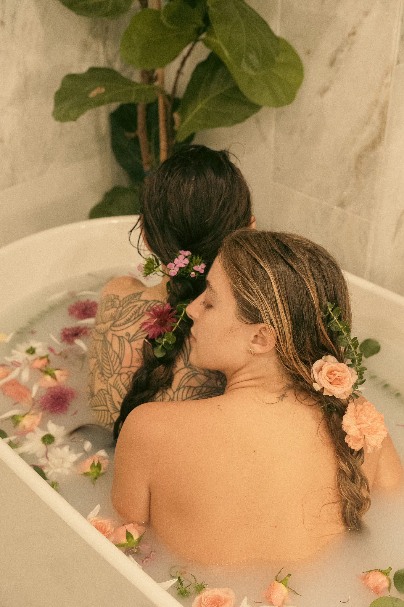 Two people in a bathtub filled with water and floating flowers, adorned with floral decorations in their hair, embracing each other.