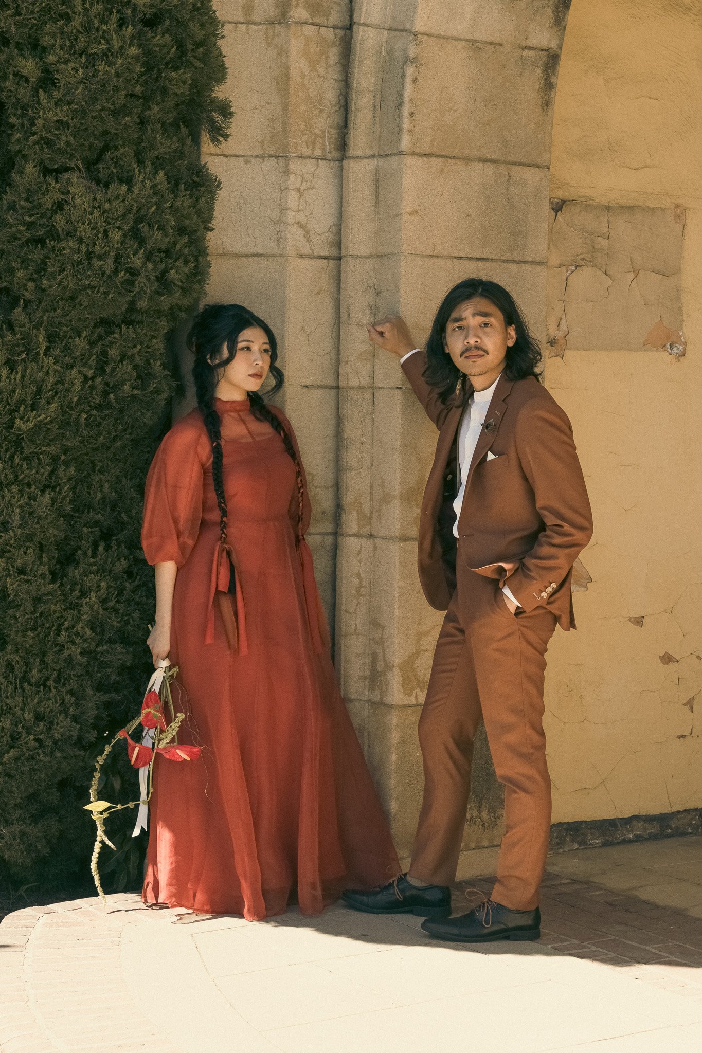 A woman in a red dress holding flowers and a man in a brown suit stand by a stone archway.