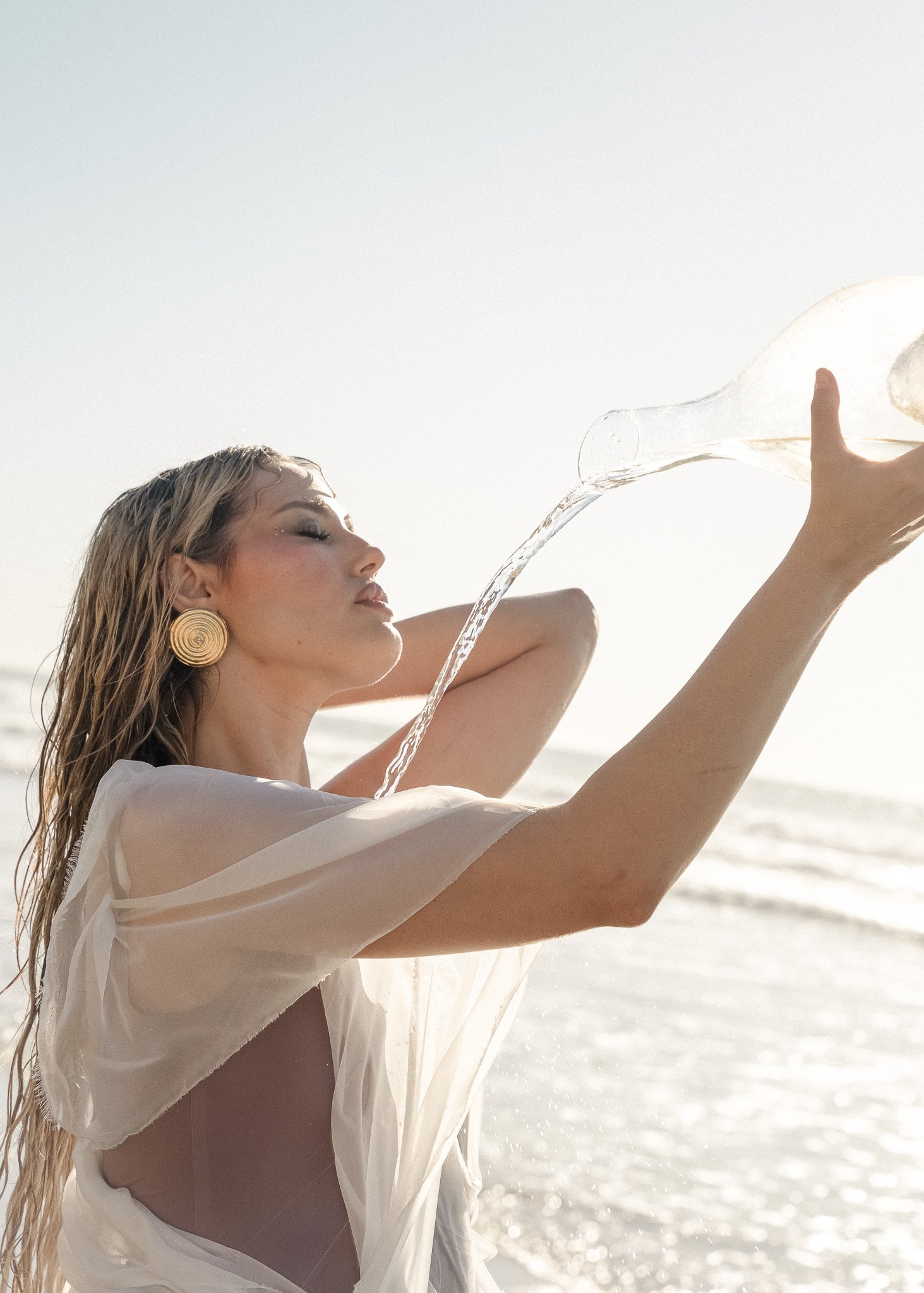 Woman holding a glass bottle pouring water over herself at the beach, wearing large spiral earrings and a white sheer garment.