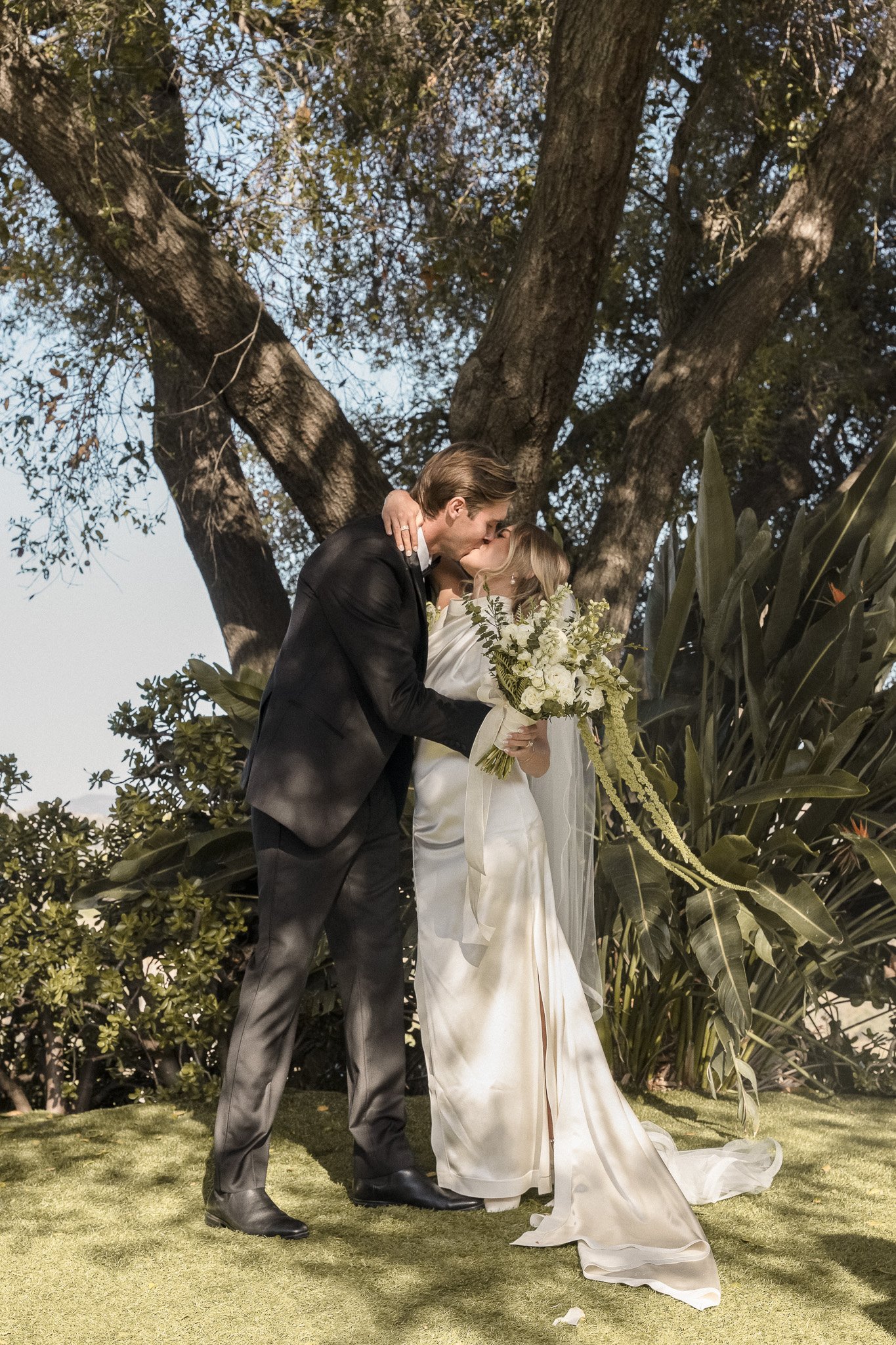 Bride and groom kissing under a tree with greenery around, bride holding a bouquet, outdoor wedding scene.