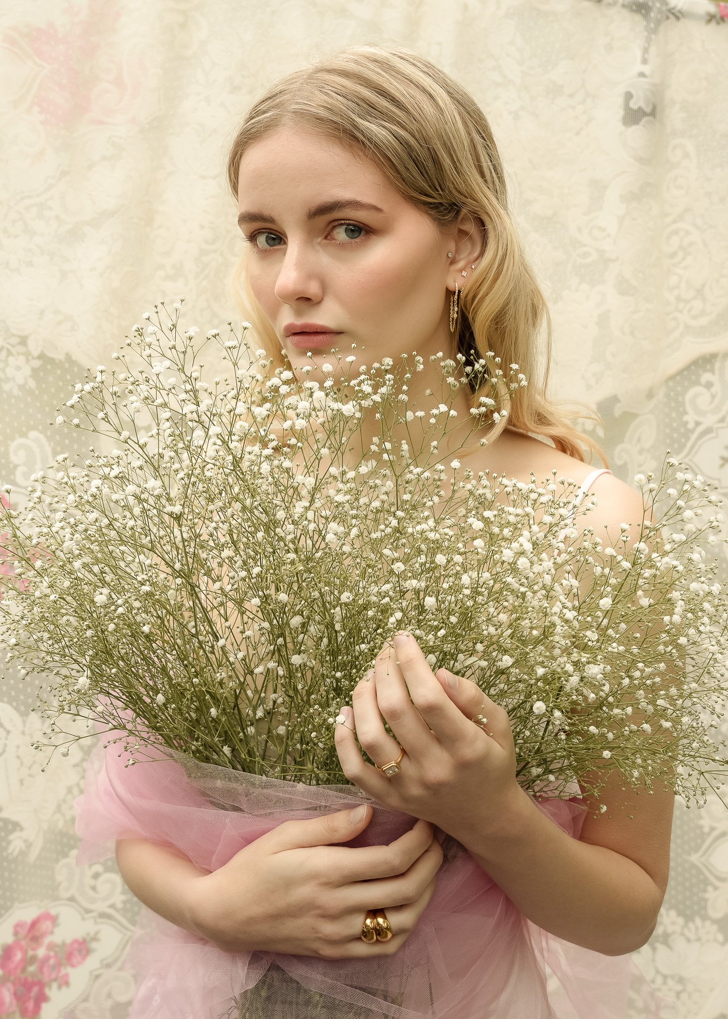 Woman holding bouquet of white baby's breath flowers, wearing gold jewelry and pink fabric, with lace curtain background.