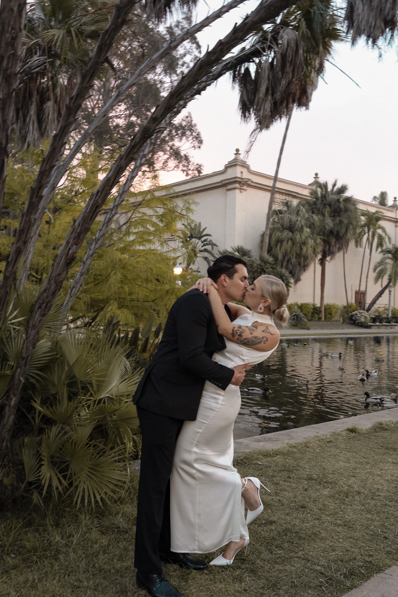 A couple in formal attire kissing near a pond surrounded by palm trees and a historic building at sunset.
