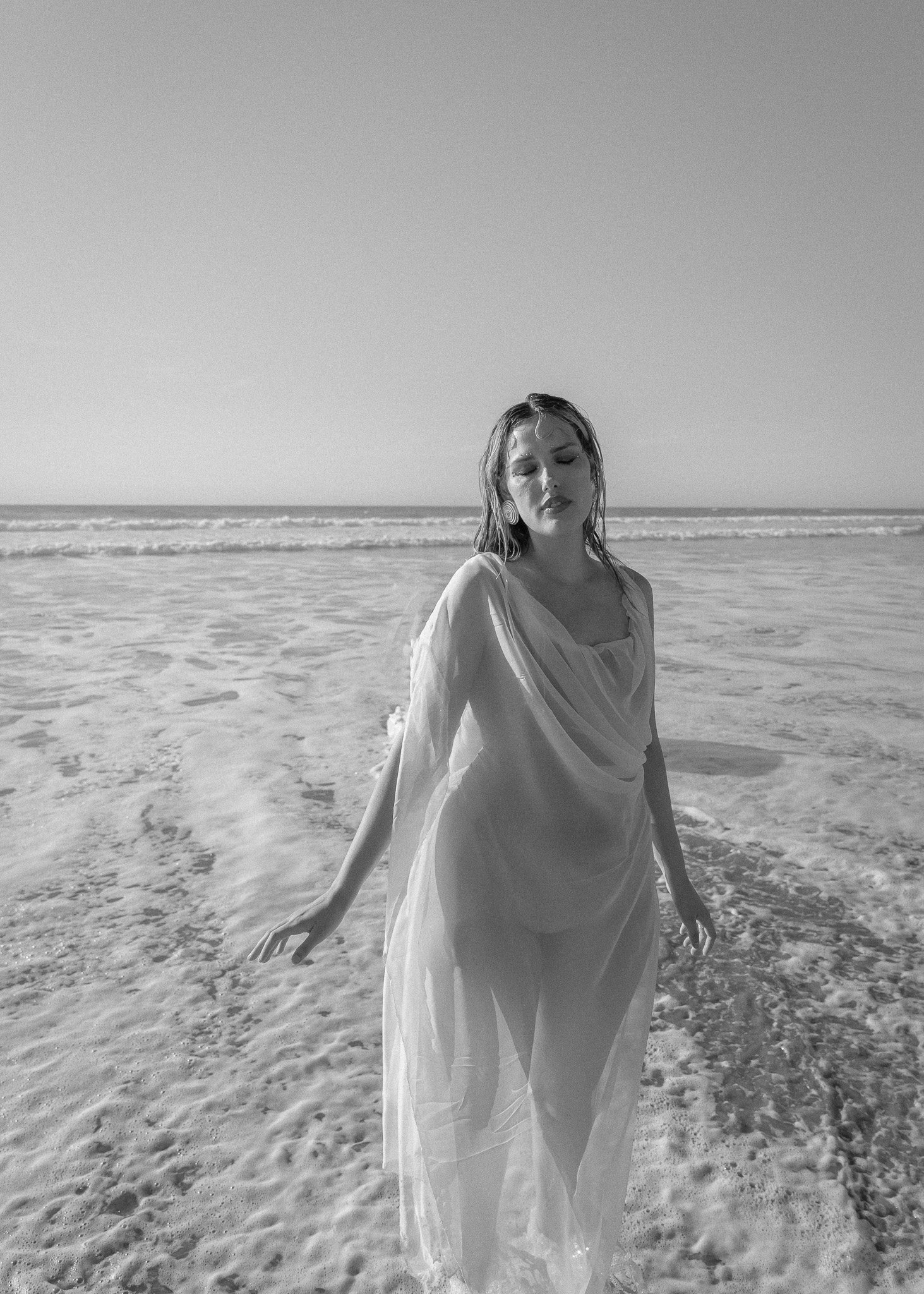 Woman in a sheer dress standing on the beach with ocean waves in the background, captured in black and white.