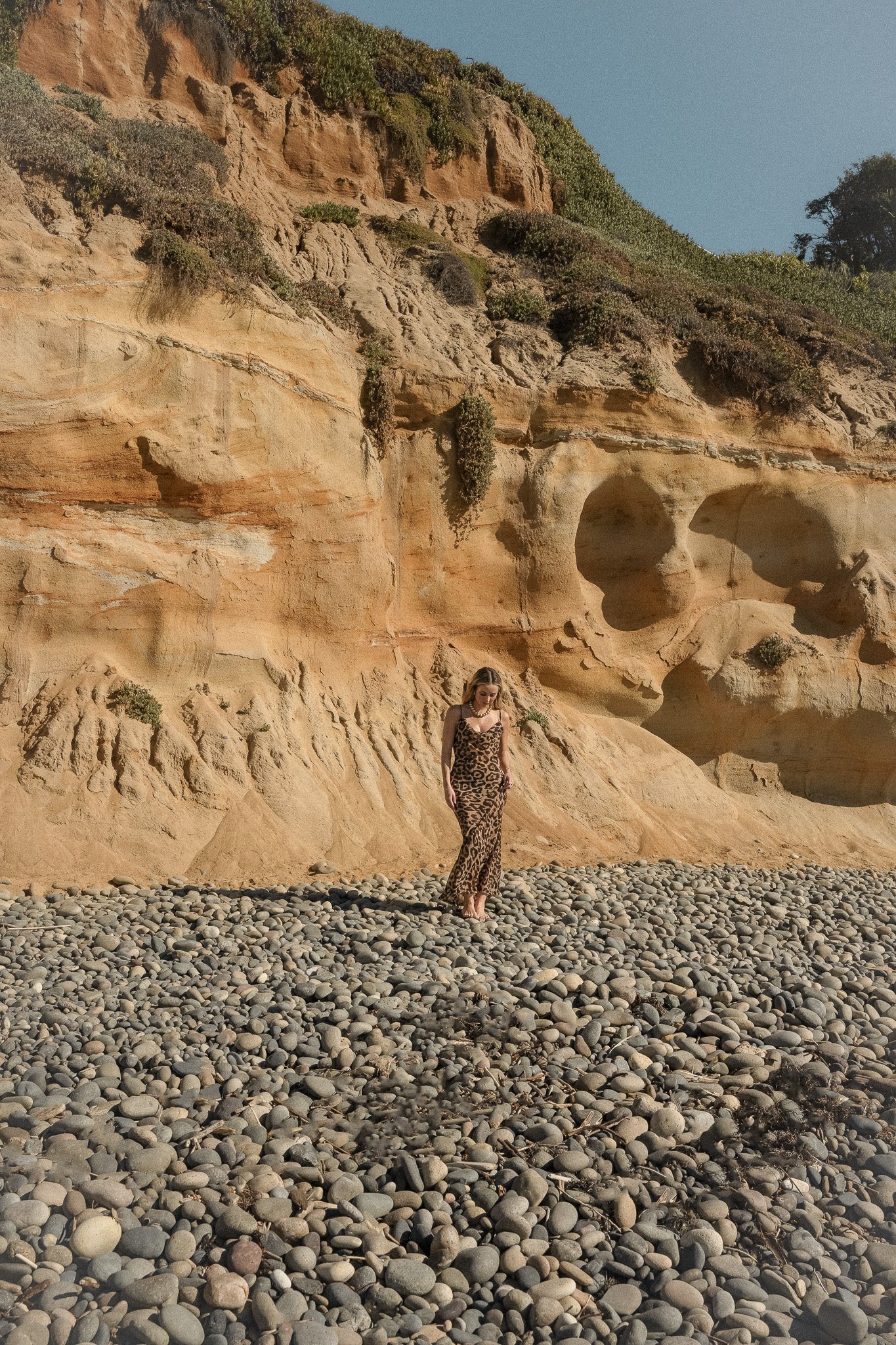 Woman in leopard print dress standing on rocky beach near sandy cliff.