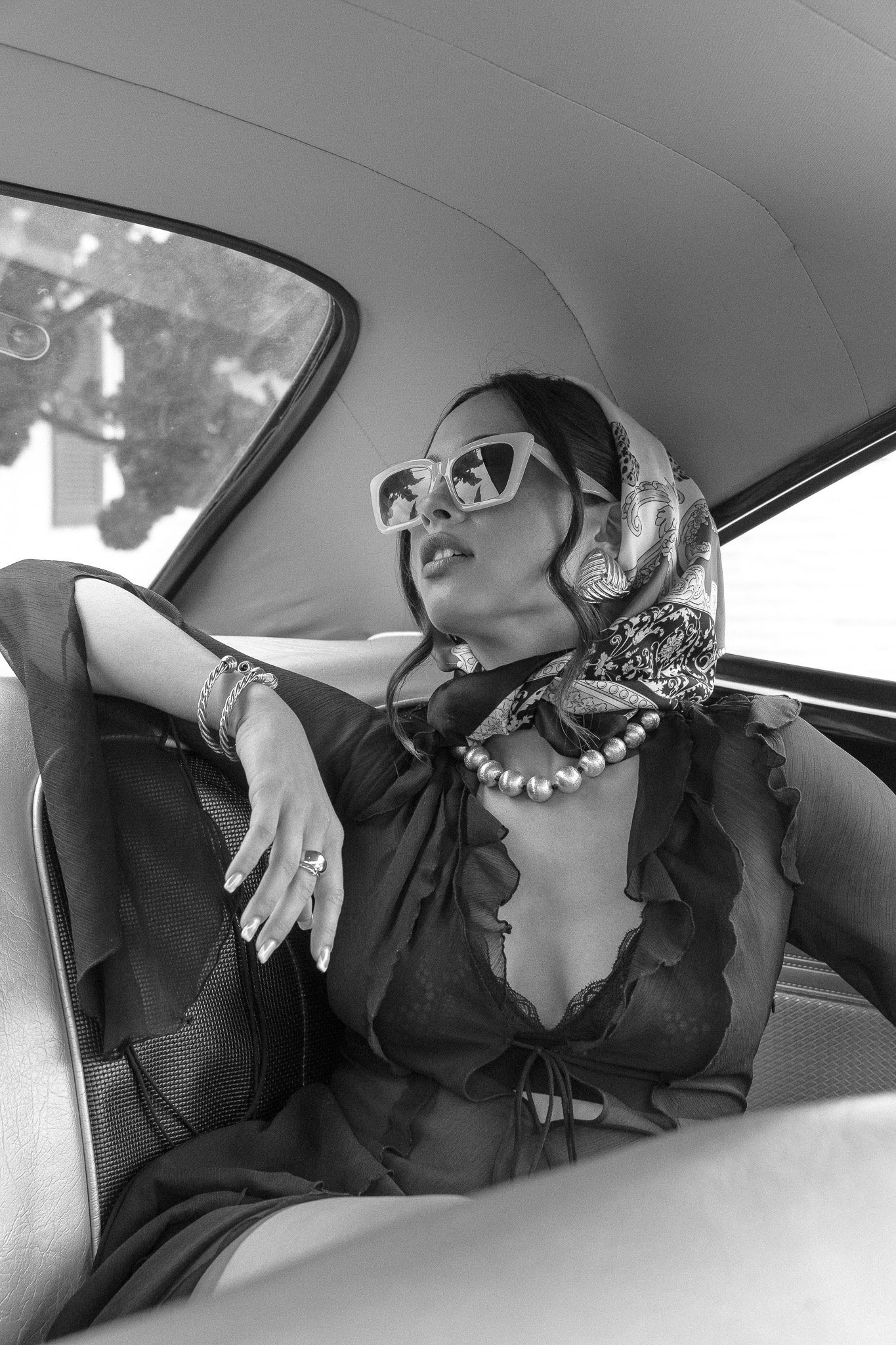 Woman in a car, wearing large sunglasses, a headscarf, and a pearl necklace, photographed in black and white.