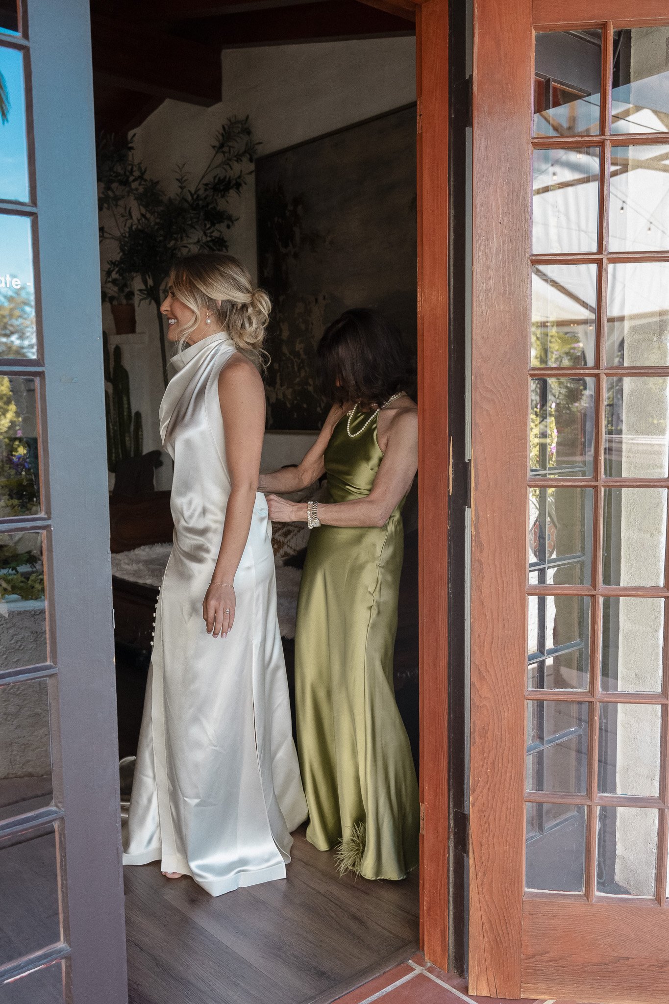 A woman in a white satin gown being assisted with her dress by another woman in a green satin gown.