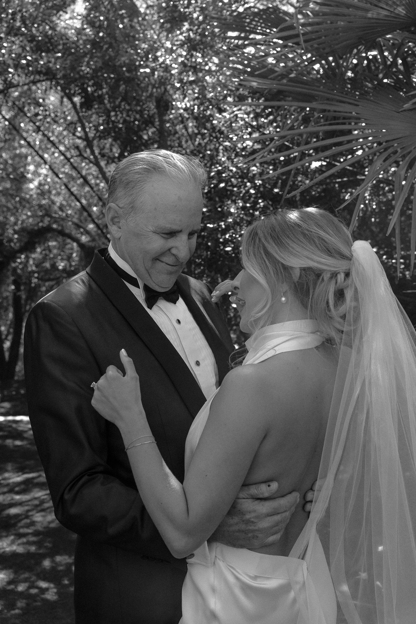 Black and white photo of a couple in formal attire; woman in a wedding dress with veil, man in a tuxedo, outdoors with foliage in the background.