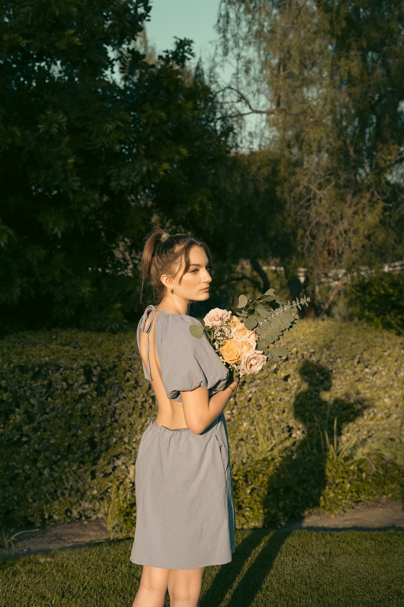 Woman in a gray dress holding a bouquet of flowers in a garden setting with trees and greenery.