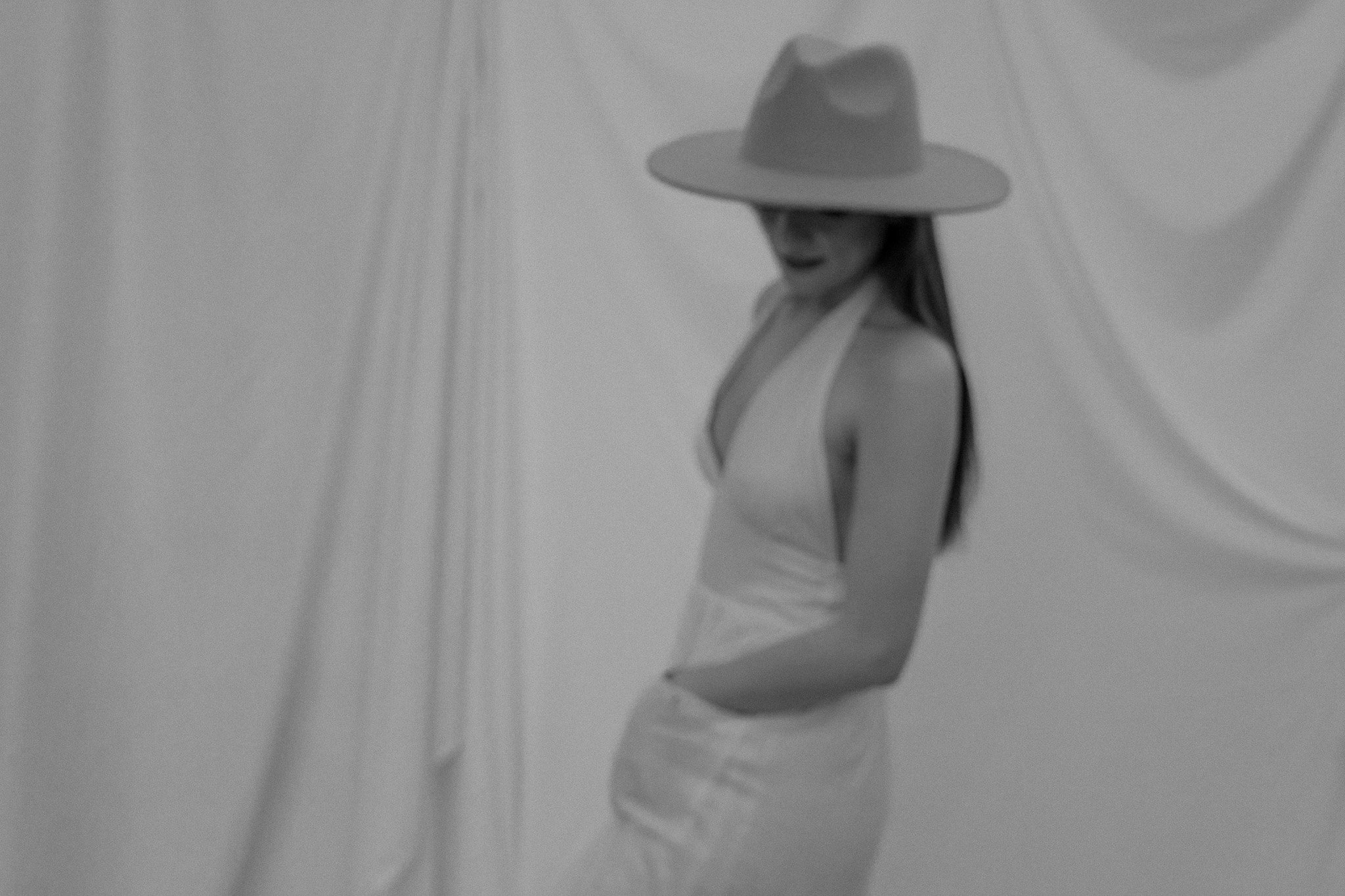 Black and white image of a woman in a dress and wide-brimmed hat, standing against a draped fabric background.