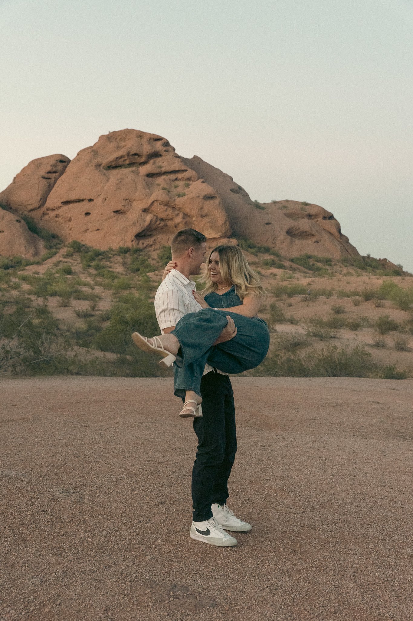 A man holding a woman in his arms in front of a rocky desert landscape.