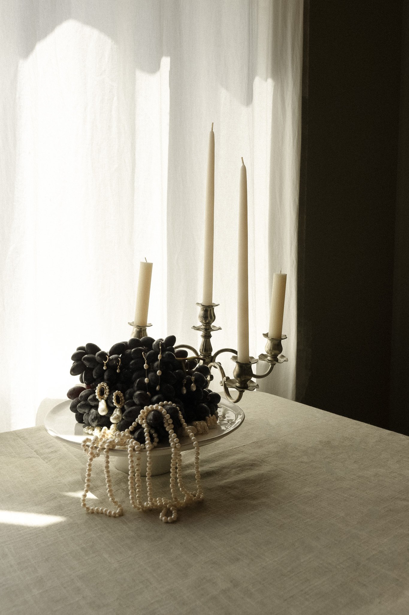 A decorative still life arrangement featuring a bowl of dark grapes adorned with pearl necklaces and earrings, next to a silver candelabra with unlit candles on a linen-covered table.