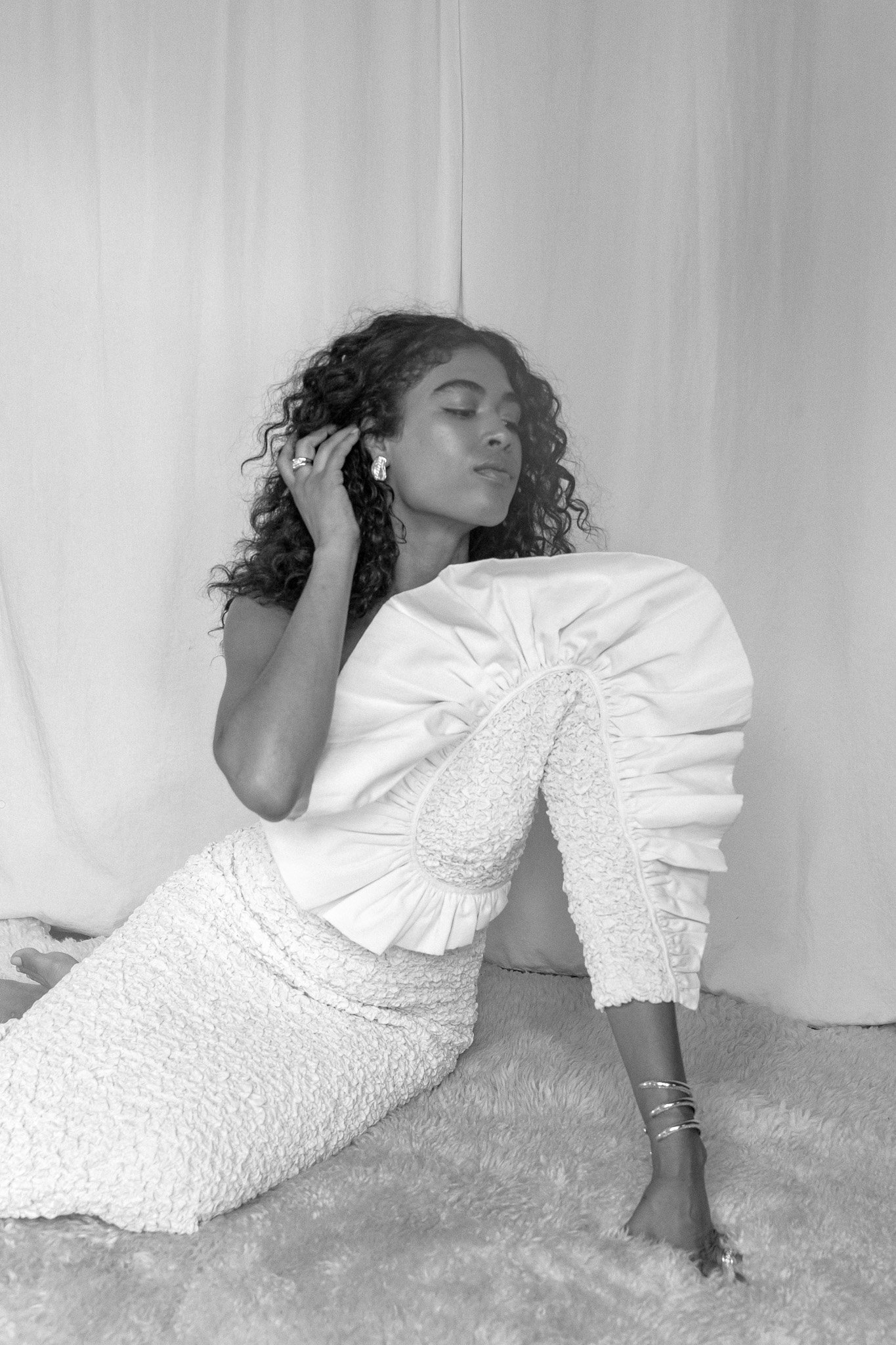 Woman in textured white outfit sitting on a fluffy surface, with one hand touching her curly hair and the other resting on her knee. The outfit features a ruffled shoulder detail.